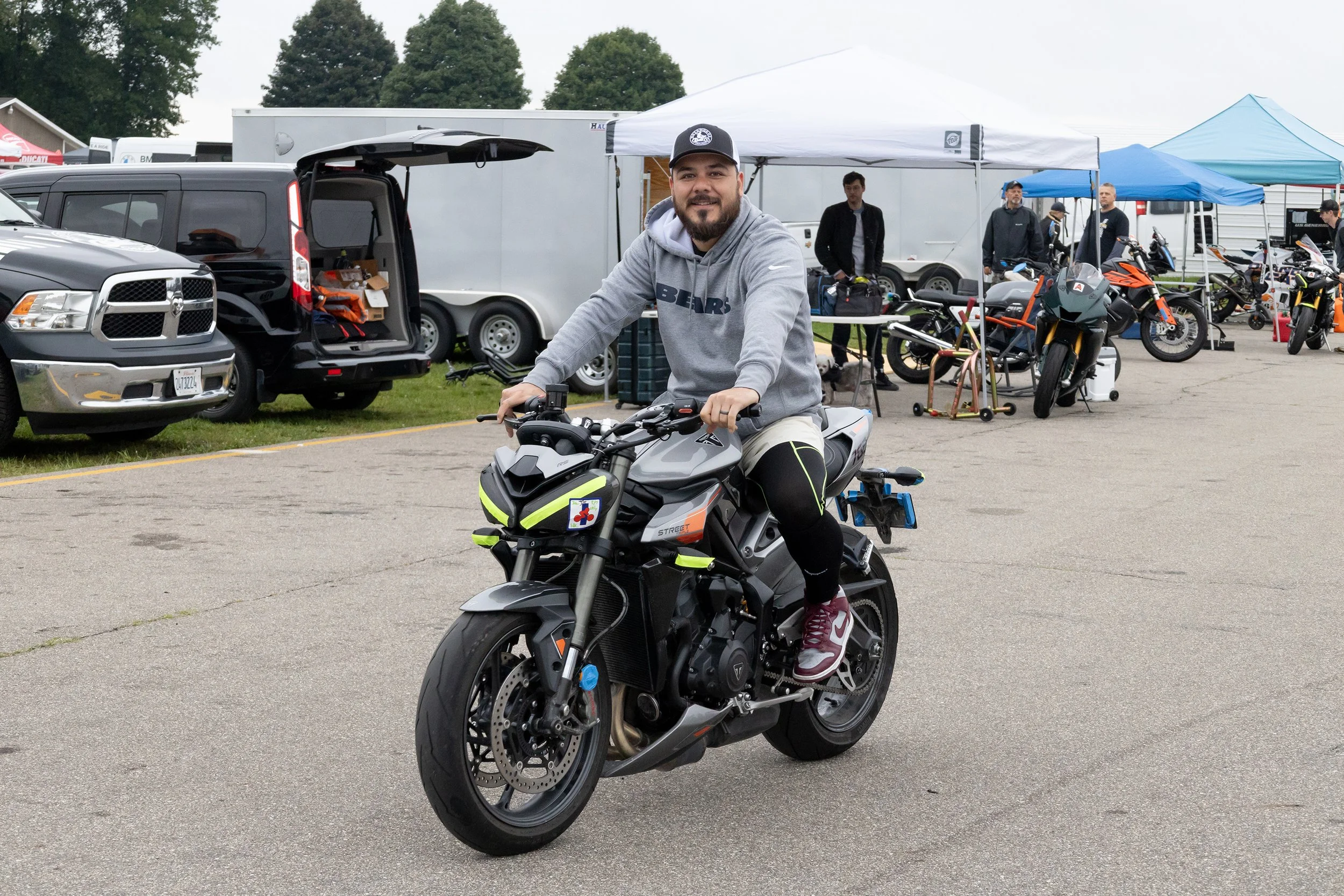 A man riding a black and gray motorcycle at an outdoor event with tents, motorcycles, and people in the background.