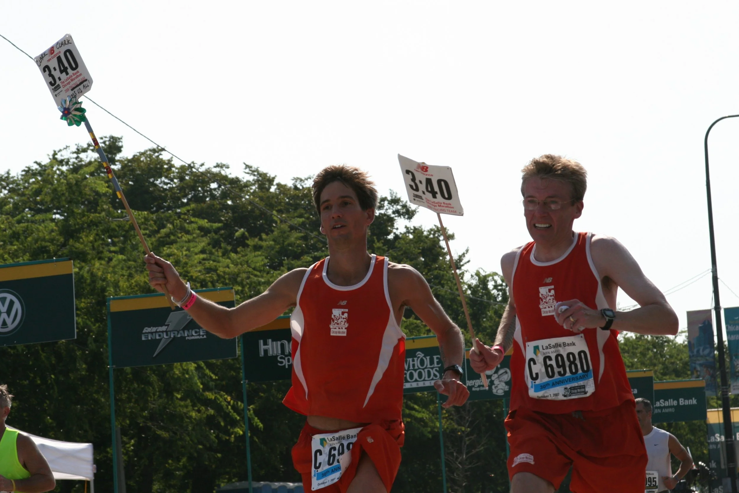 Two male marathon runners in red uniforms running during a race, holding a sign with the number 3:40, with trees and race banners in the background.