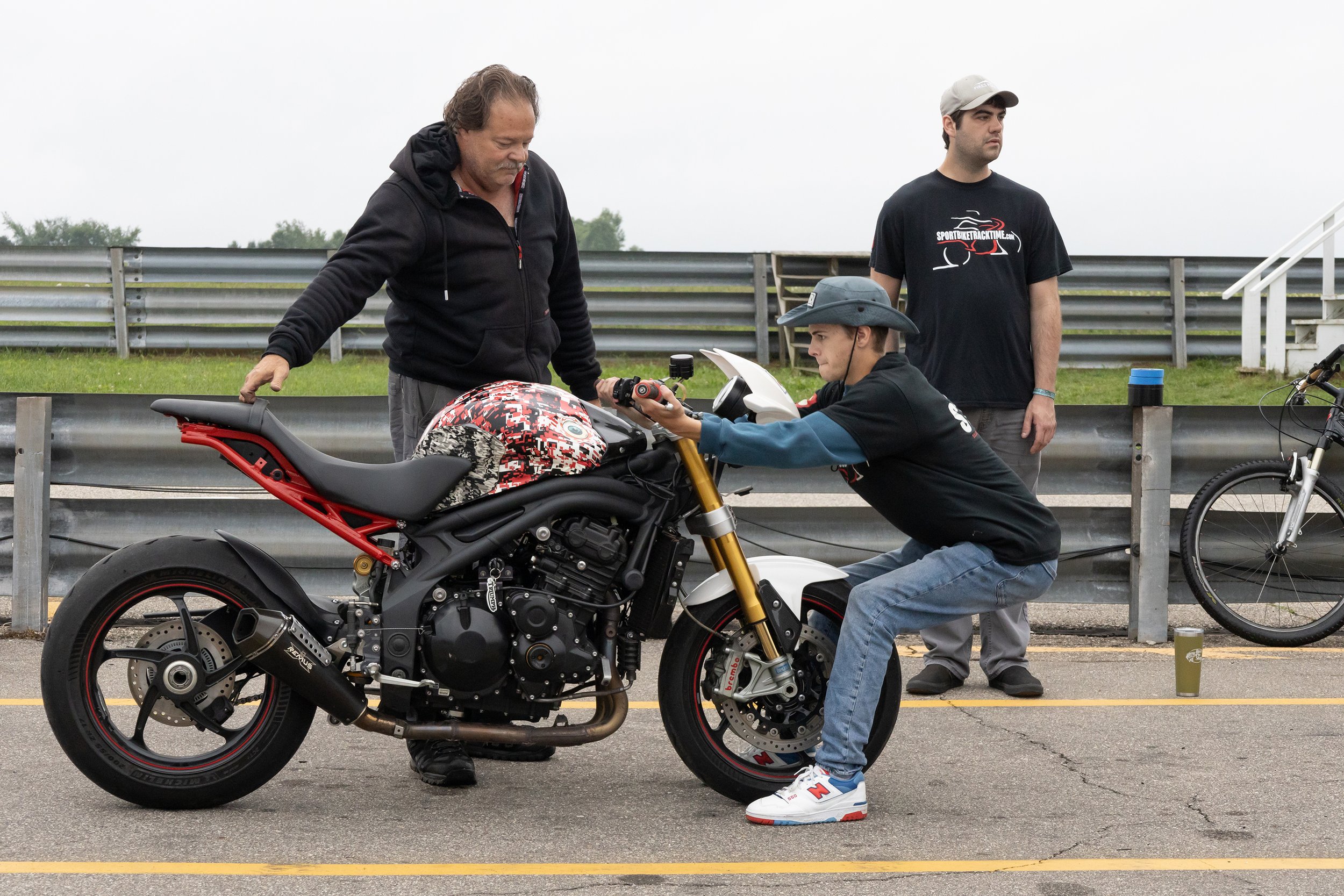 Three men are at a race track with a motorcycle. One man is crouched down, adjusting something on the motorcycle. The second man, taller, stands nearby watching, wearing a black t-shirt. The third man is looking down at the bike, wearing a black hood