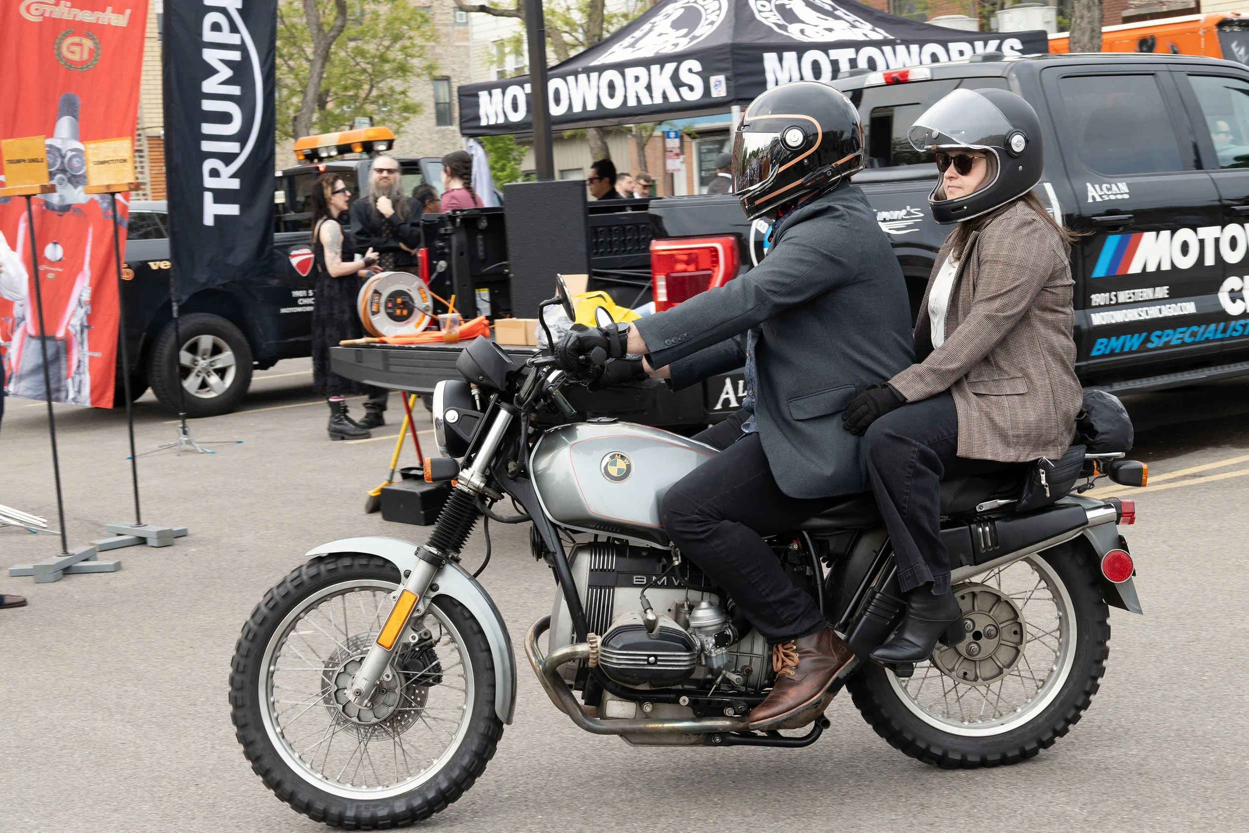 A person riding a BMW motorcycle at an outdoor event, wearing a helmet and glasses, with a woman sitting behind them on the motorcycle, both dressed warmly. In the background, there are tents, banners, and people gathered around booths.