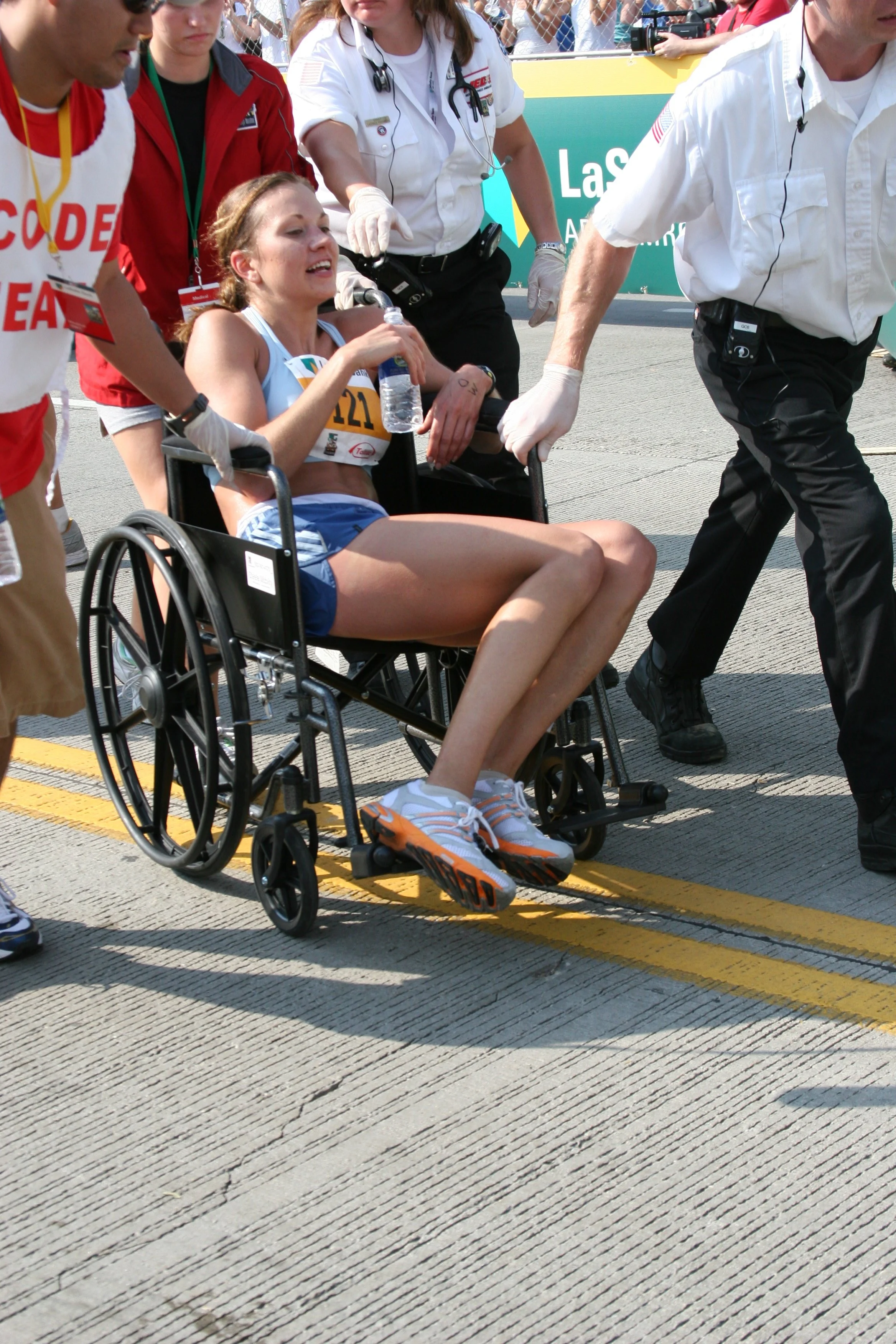 A female athlete in a wheelchair being assisted by medical personnel during a race, holding a water bottle and smiling, with a crowd and event banners in the background.