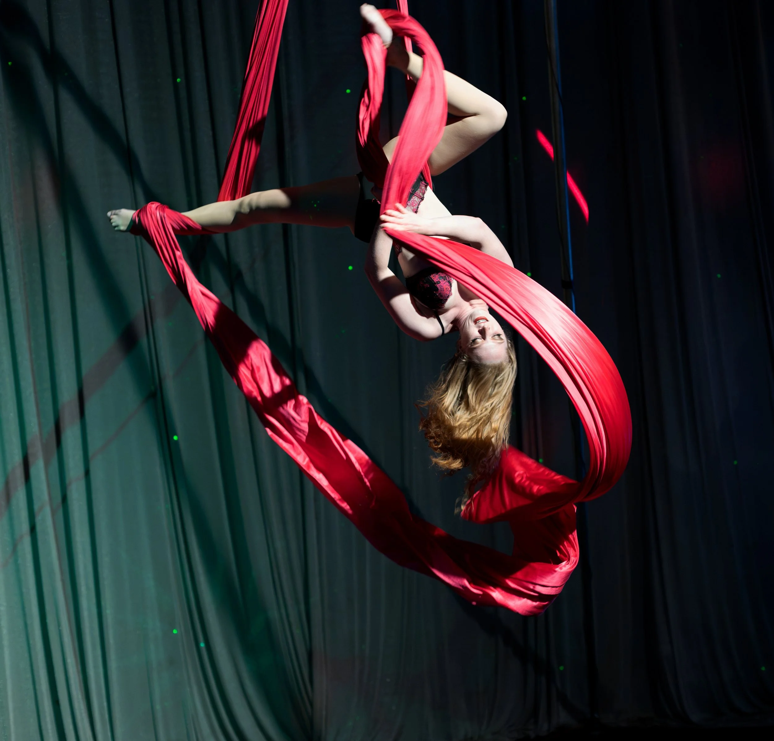 A female aerial silk performer hanging upside down on red silk fabric during a performance on stage with black curtains in the background.