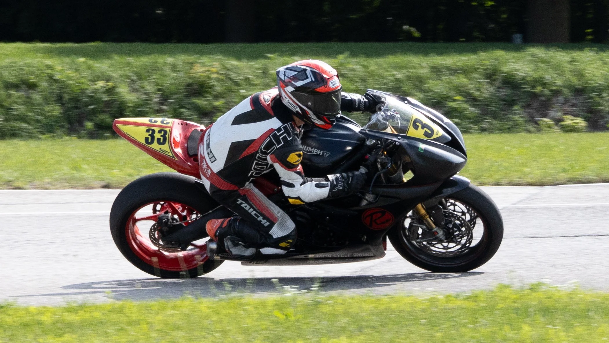 A motorcycle racer in a black, red, and yellow racing suit and helmet riding a black motorcycle with yellow and red accents on a race track with green grass and bushes in the background.