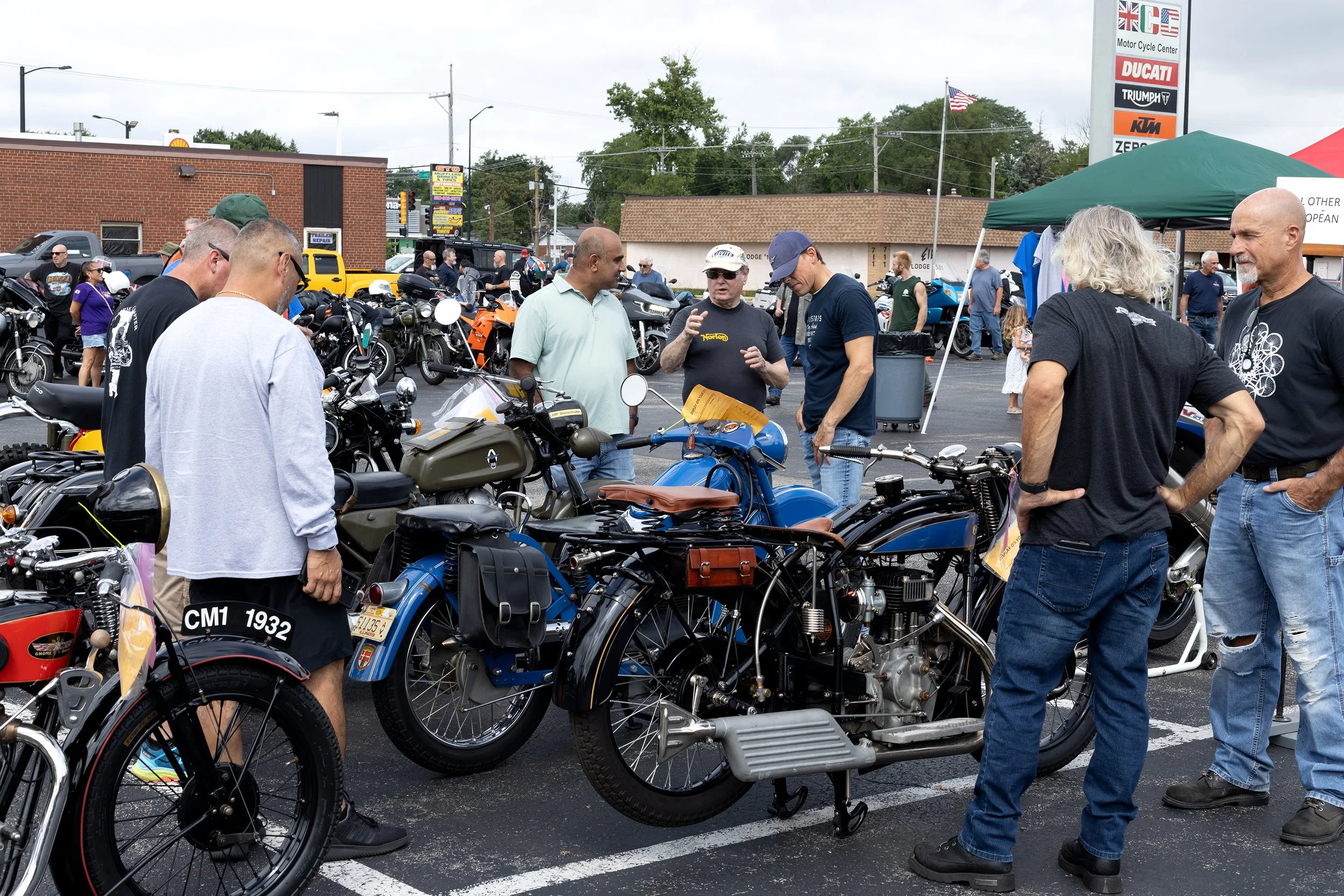 People gather around vintage and modern motorcycles at a motorcycle show in a parking lot, with a tent and a tall sign in the background.