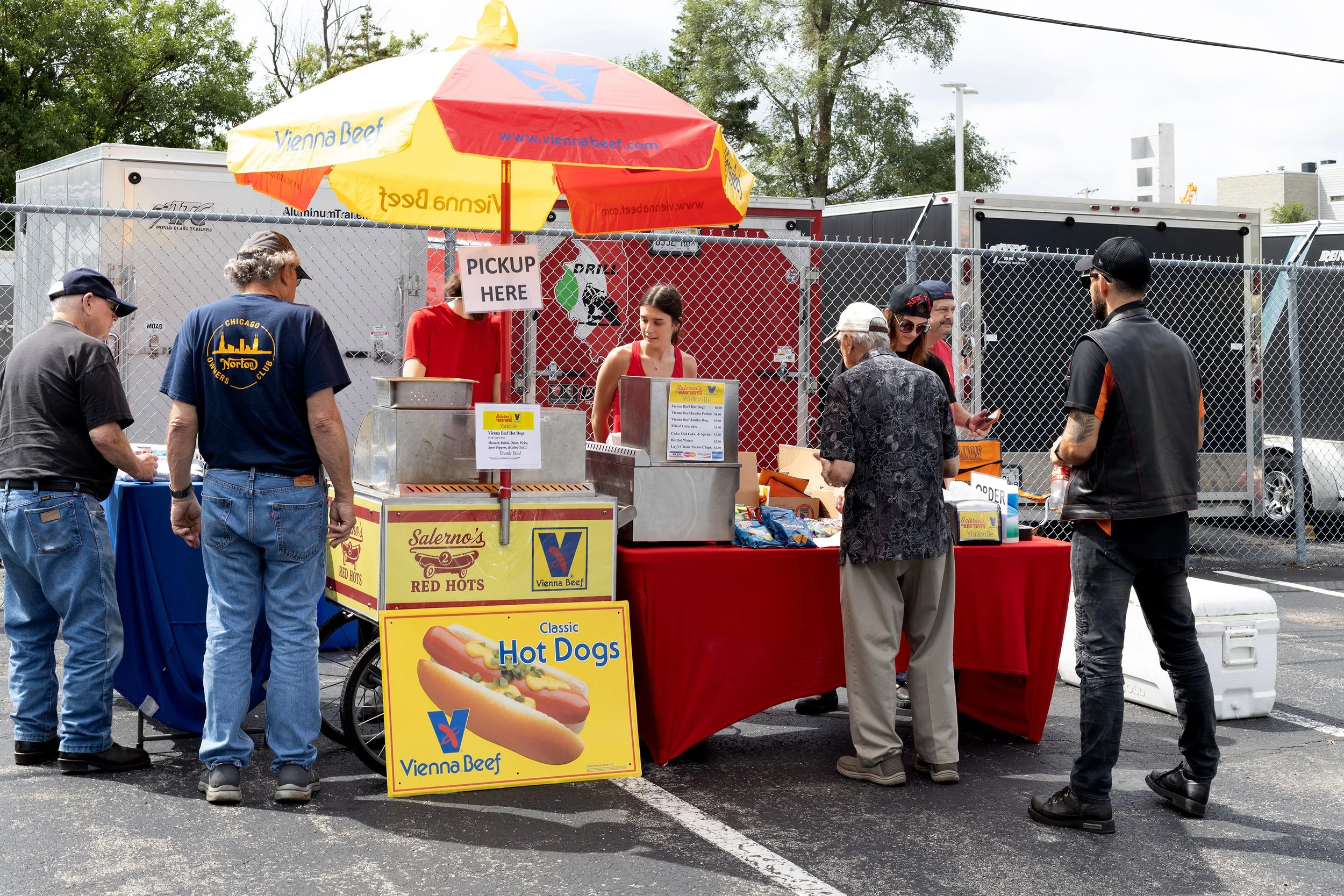 People ordering food from a hot dog stand at an outdoor event, with a yellow and red umbrella and a sign indicating 