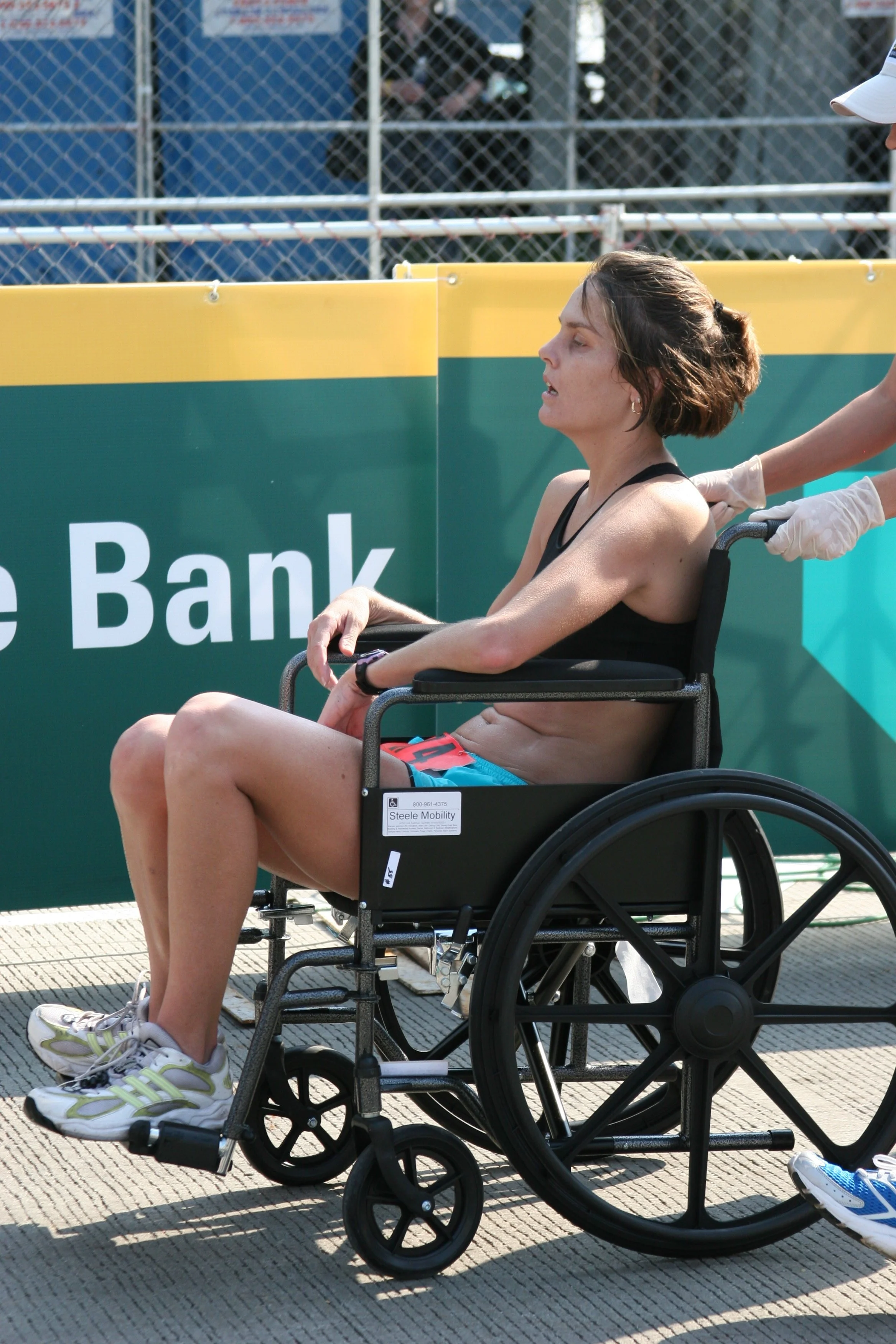 A woman in a wheelchair being pushed by a person wearing gloves on a sports field. The woman has short brown hair and is dressed in athletic attire. There are fences and advertisements in the background.
