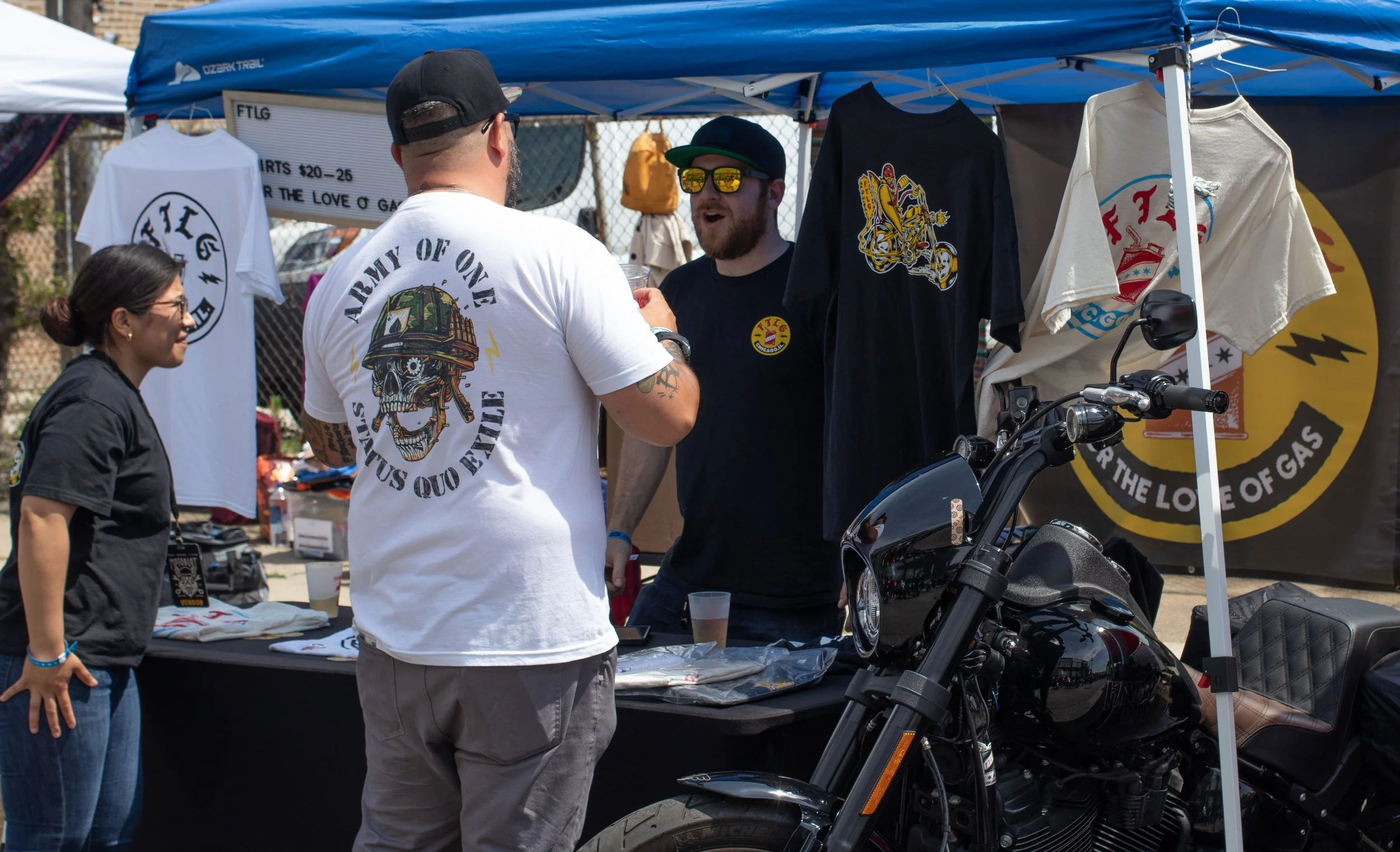 People at an outdoor market or event booth selling or displaying motorcycle-themed clothing and accessories, including T-shirts with skull and motorcycle designs, under a blue canopy.