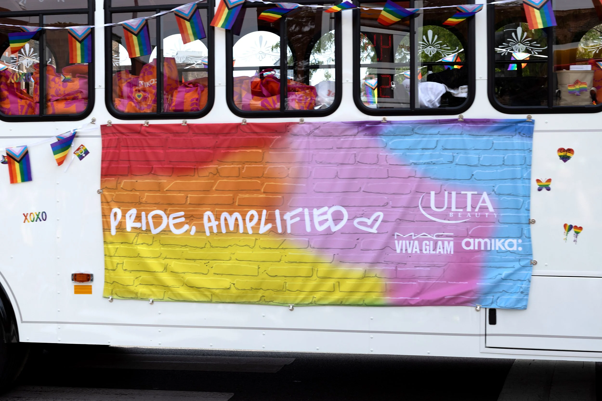 A decorated vehicle with rainbow pride flags and a large colorful banner displaying the words "Pride, Amplified" along with logos of various beauty and cosmetic brands, a small rainbow heart sticker, and butterfly and heart stickers featuring rainbow