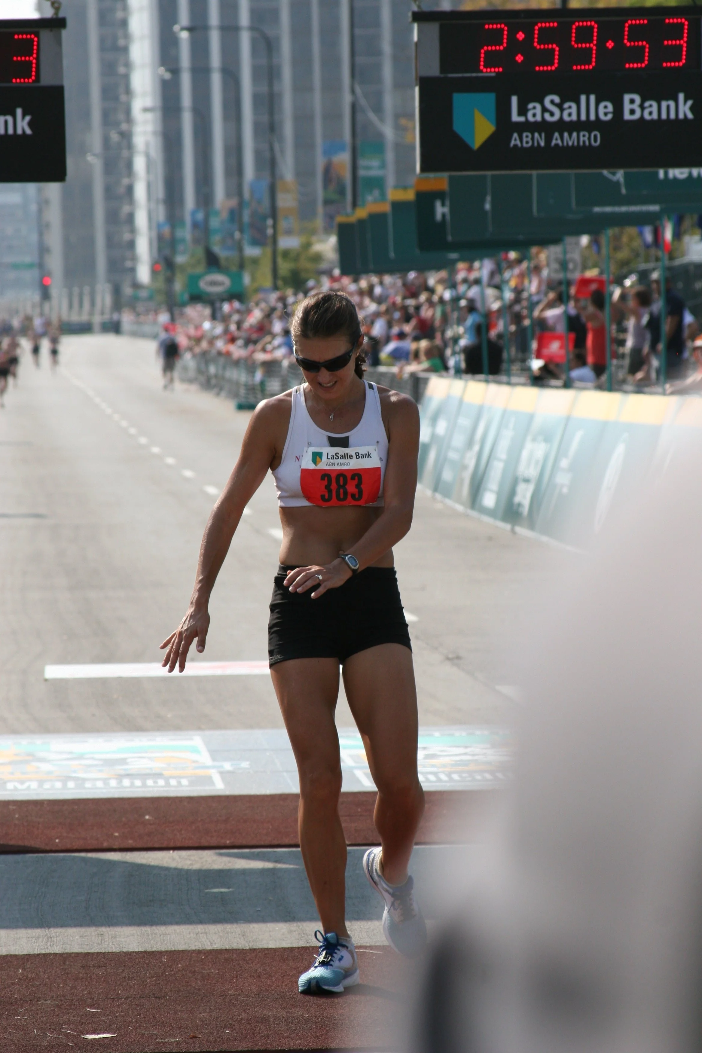 Female marathon runner crossing the finish line during a race, wearing a white tank top with bib number 383, black shorts, and white running shoes, in an urban city setting with spectators and digital clock displaying 2:59:53.