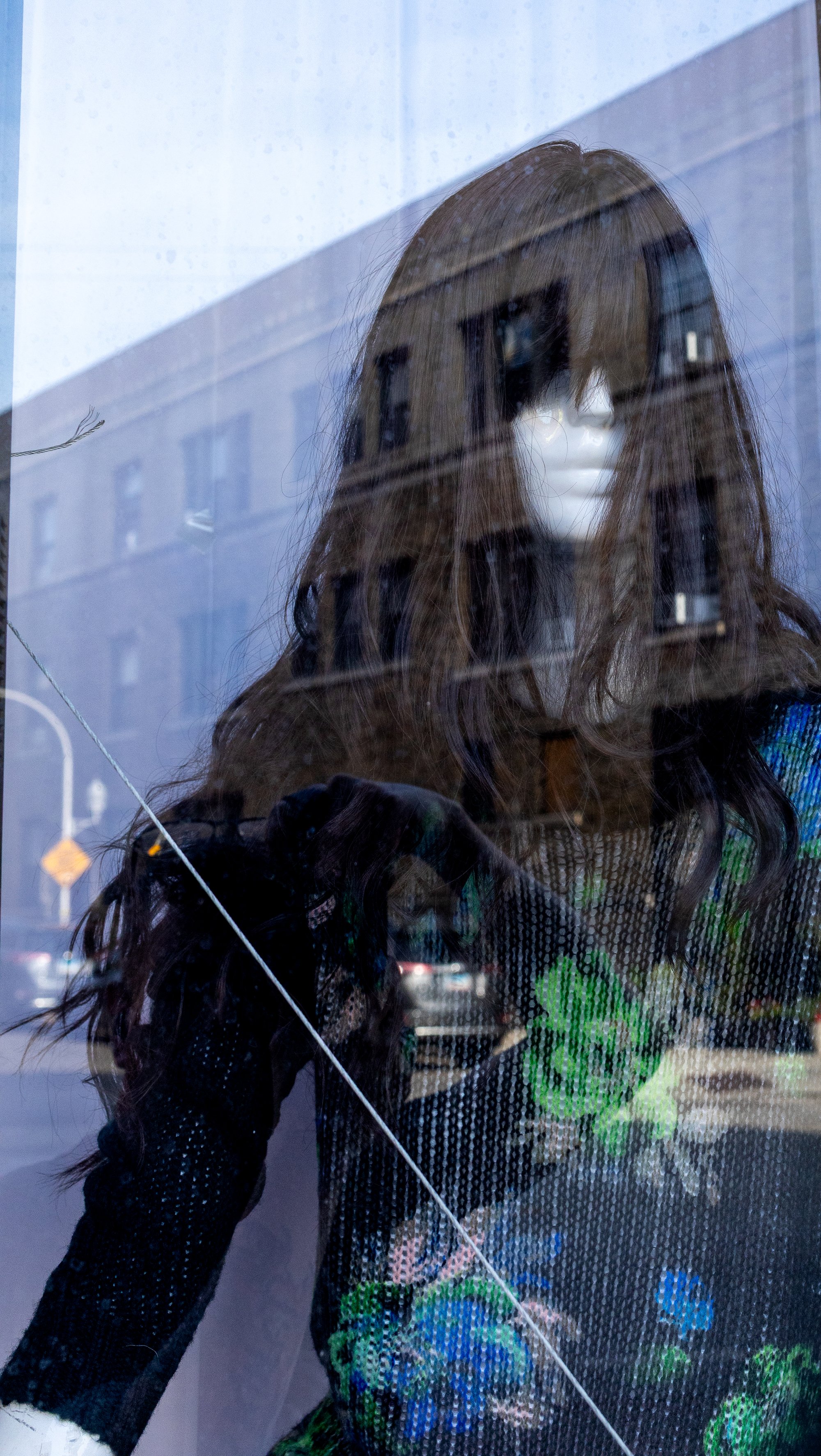 Reflection of a mannequin with long hair and a face mask in a store window, with a building and street visible in the background.