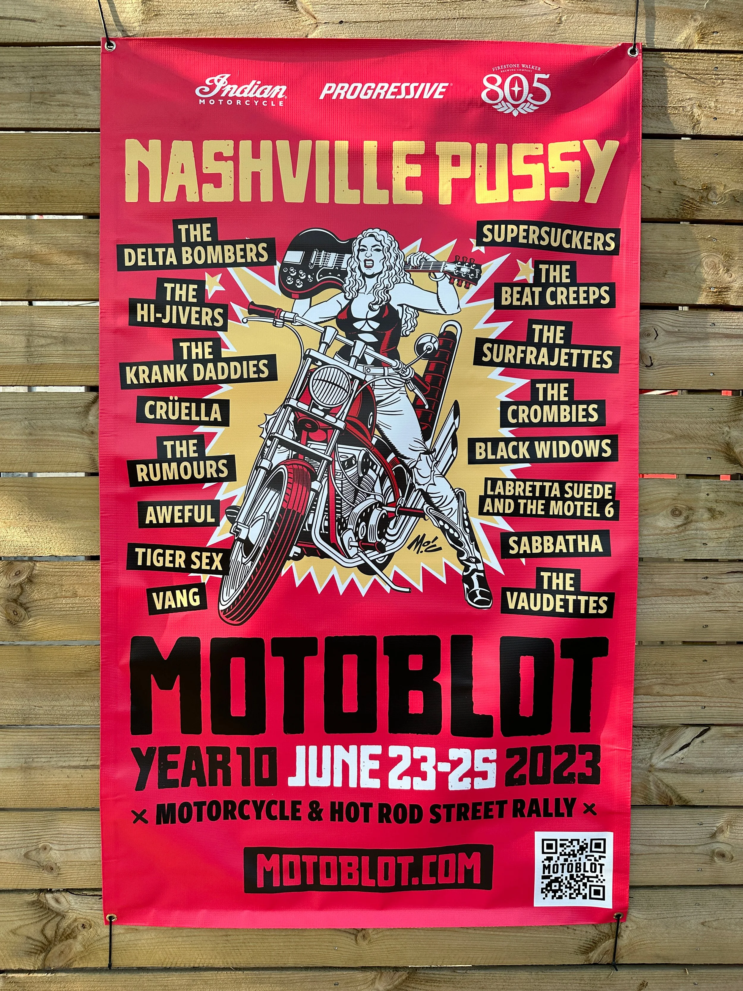 Pink event banner announcing Nashville Pussey, a motorcycle and hot rod street rally from June 23 to 25, 2023, with a central illustration of a woman riding a motorcycle, holding a guitar, surrounded by various band names and logos, mounted on a wood
