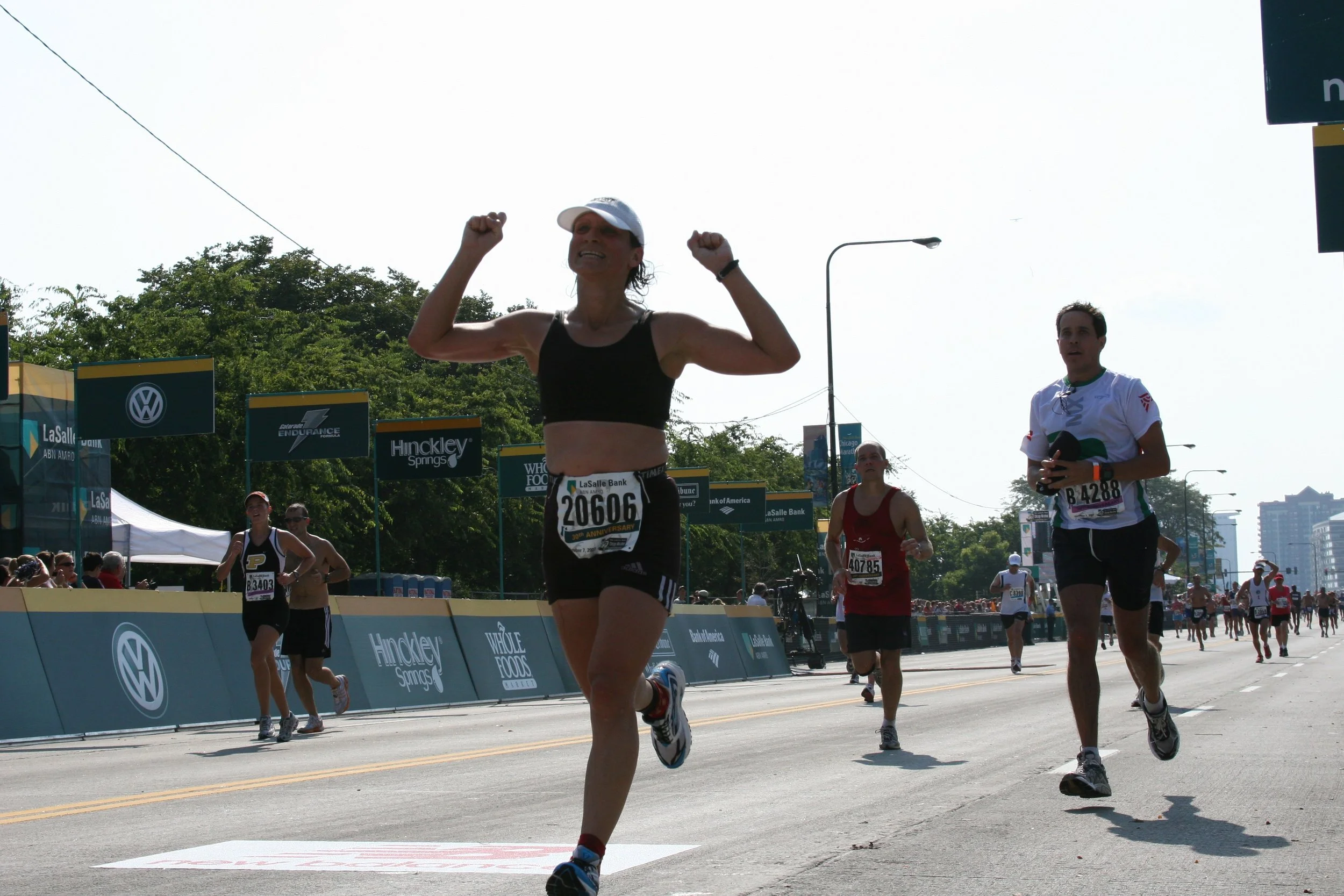 A group of marathon runners competing on a city street during a race. The runner in the foreground is a woman with a white cap and black sports bra, smiling and flexing her arms as she runs. Other runners and spectators are visible in the background,