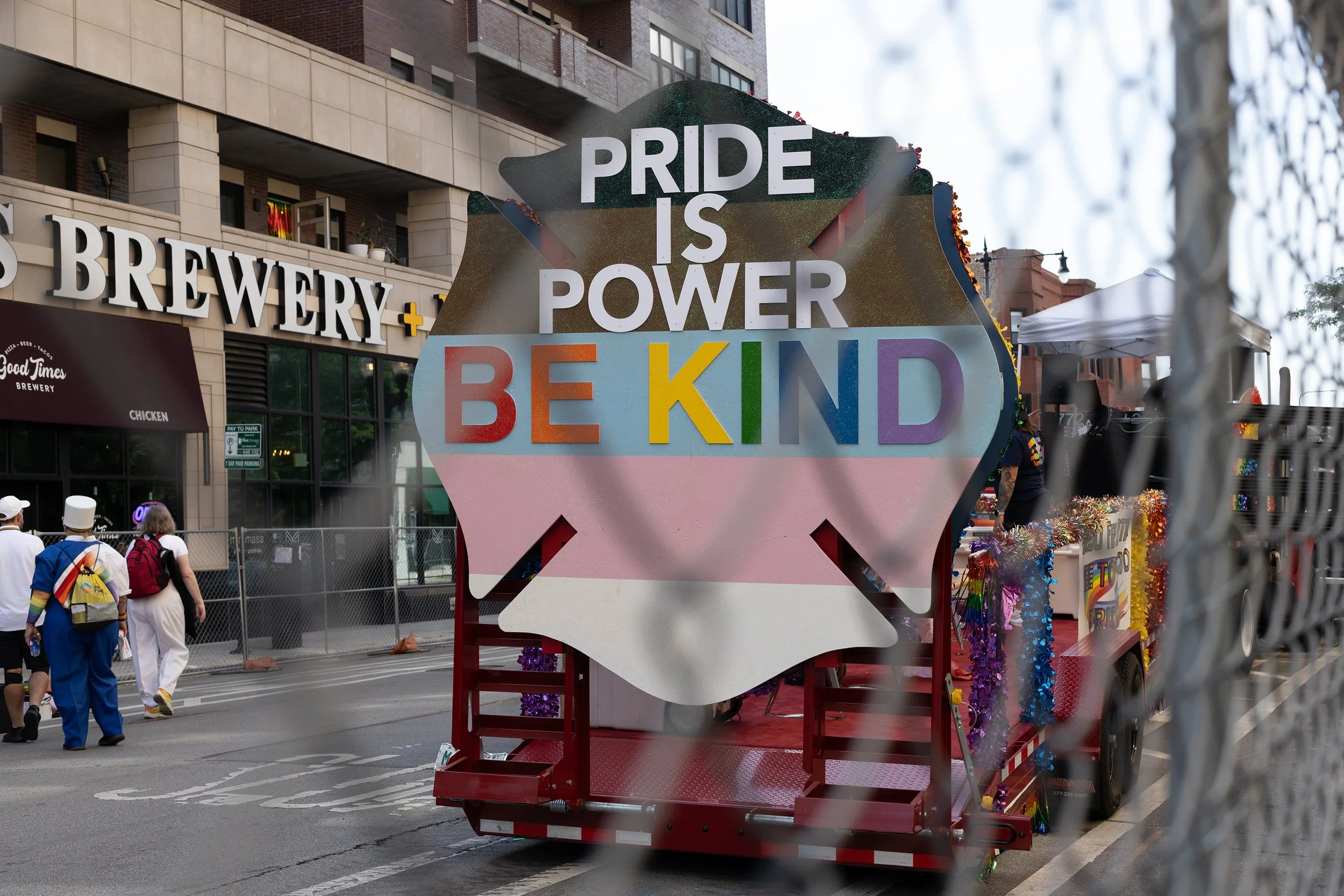 A decorated float at a parade with a sign that reads "PRIDE IS POWER BE KIND." The float is on a city street, with people walking nearby, and a metal fence partially in view.