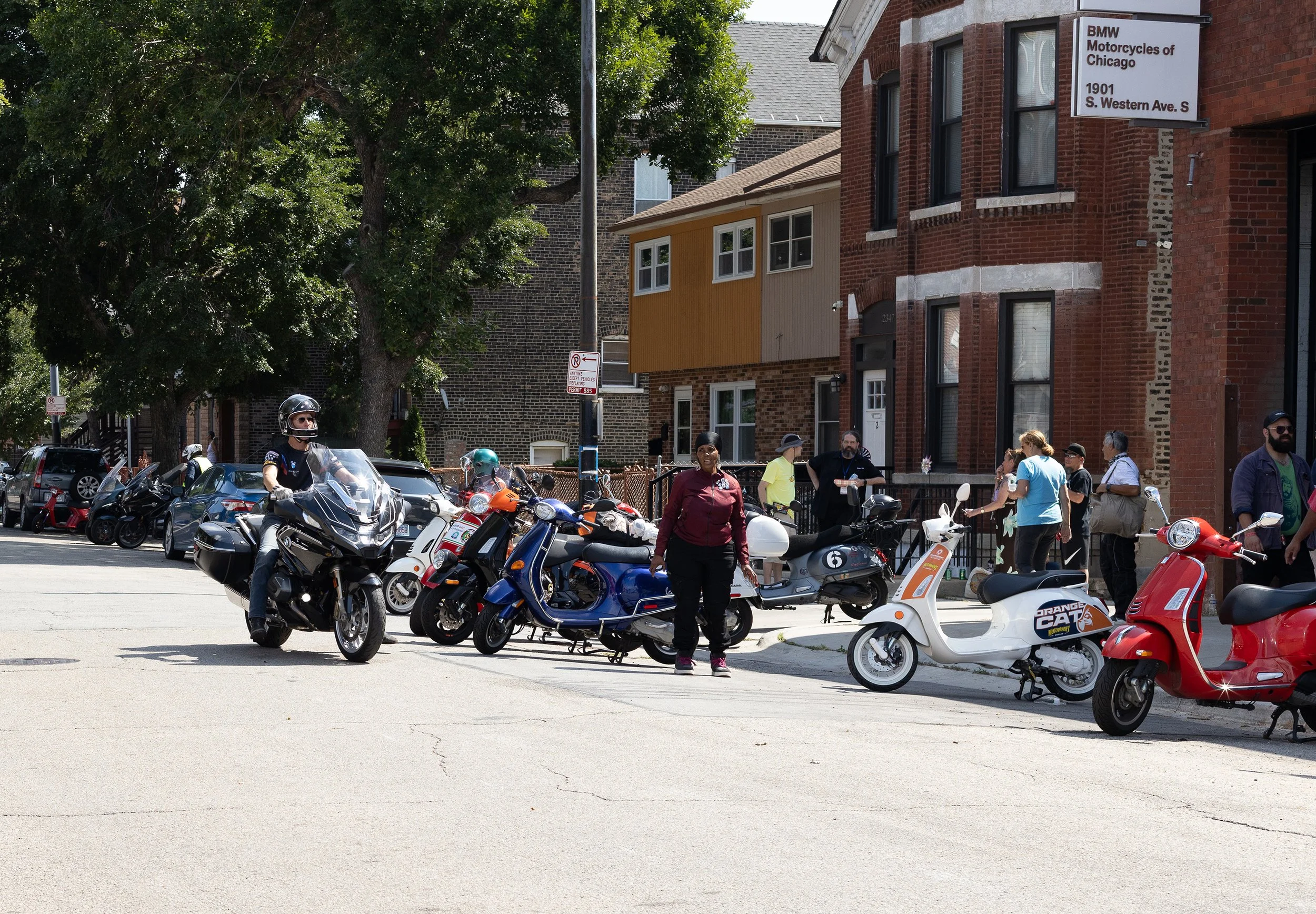 People standing and walking near parked motorcycles and scooters on a city street, with a brick building and a sign for BMW Motorcycles of Chicago in the background.