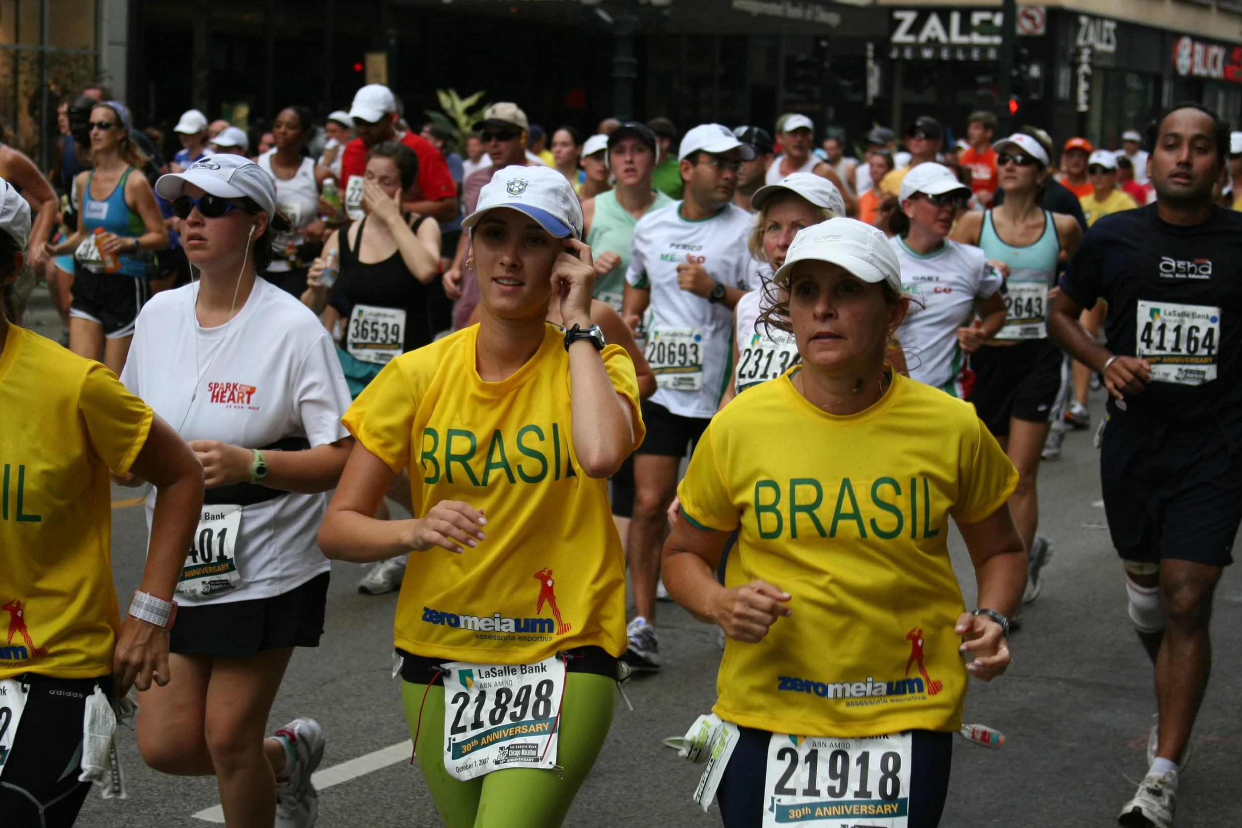 A diverse group of marathon runners participating in a race, wearing athletic clothing and race bibs, some with hats and sunglasses, running on city streets.