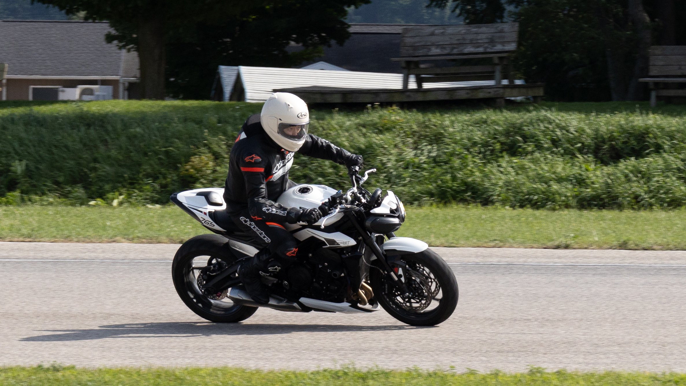 A person riding a black and white sport motorcycle on a paved road, wearing a white helmet and black racing gear with red and white accents, grass and trees in the background.