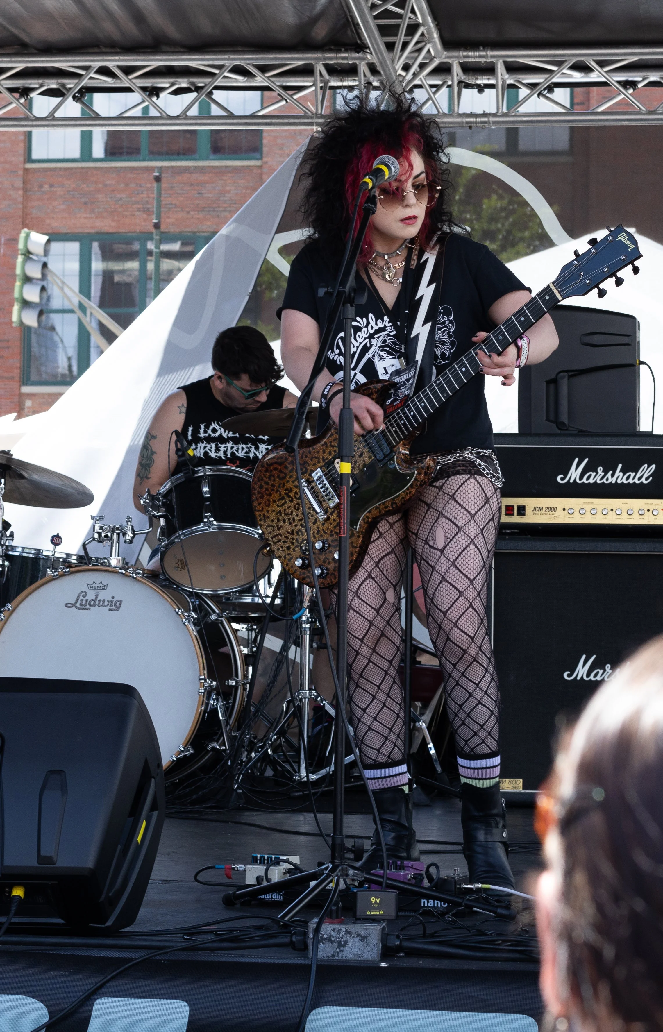 A woman with curly black and pink hair playing an electric guitar on stage, with a drummer in the background and musical equipment around, including a Marshall amplifier and drum set.