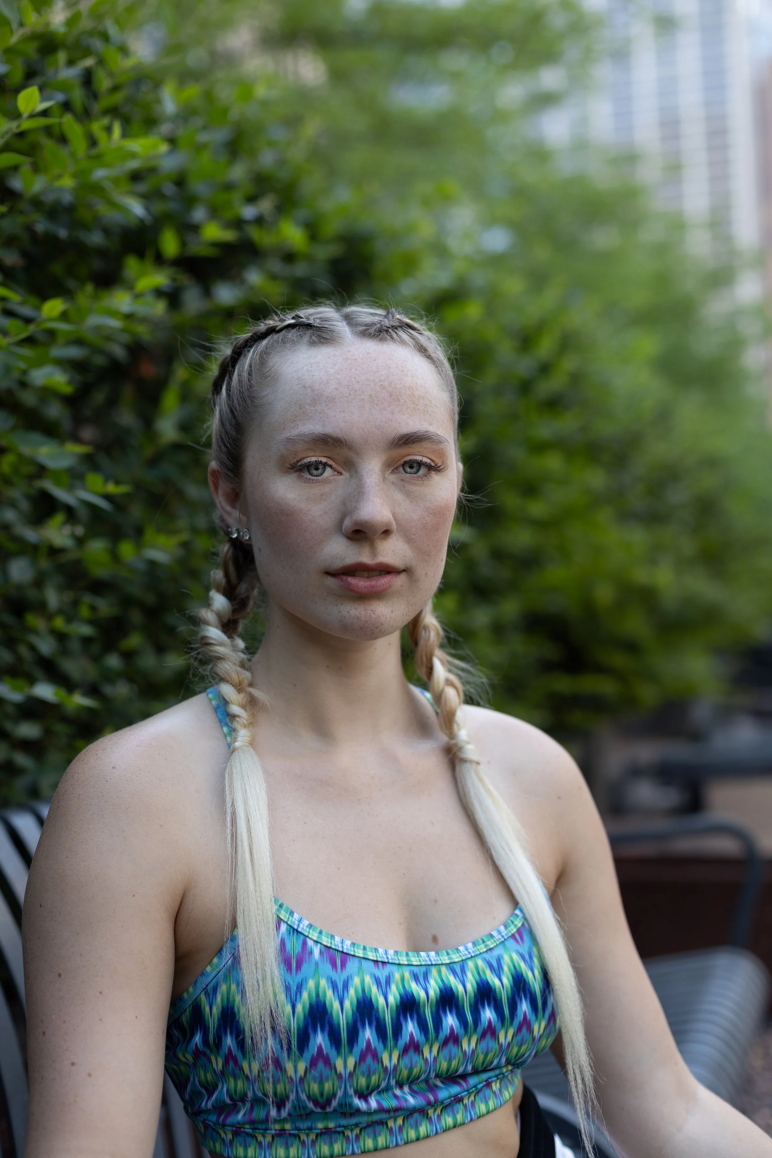 A young woman with braided hair and light skin standing outdoors in front of green bushes, wearing a colorful tank top.