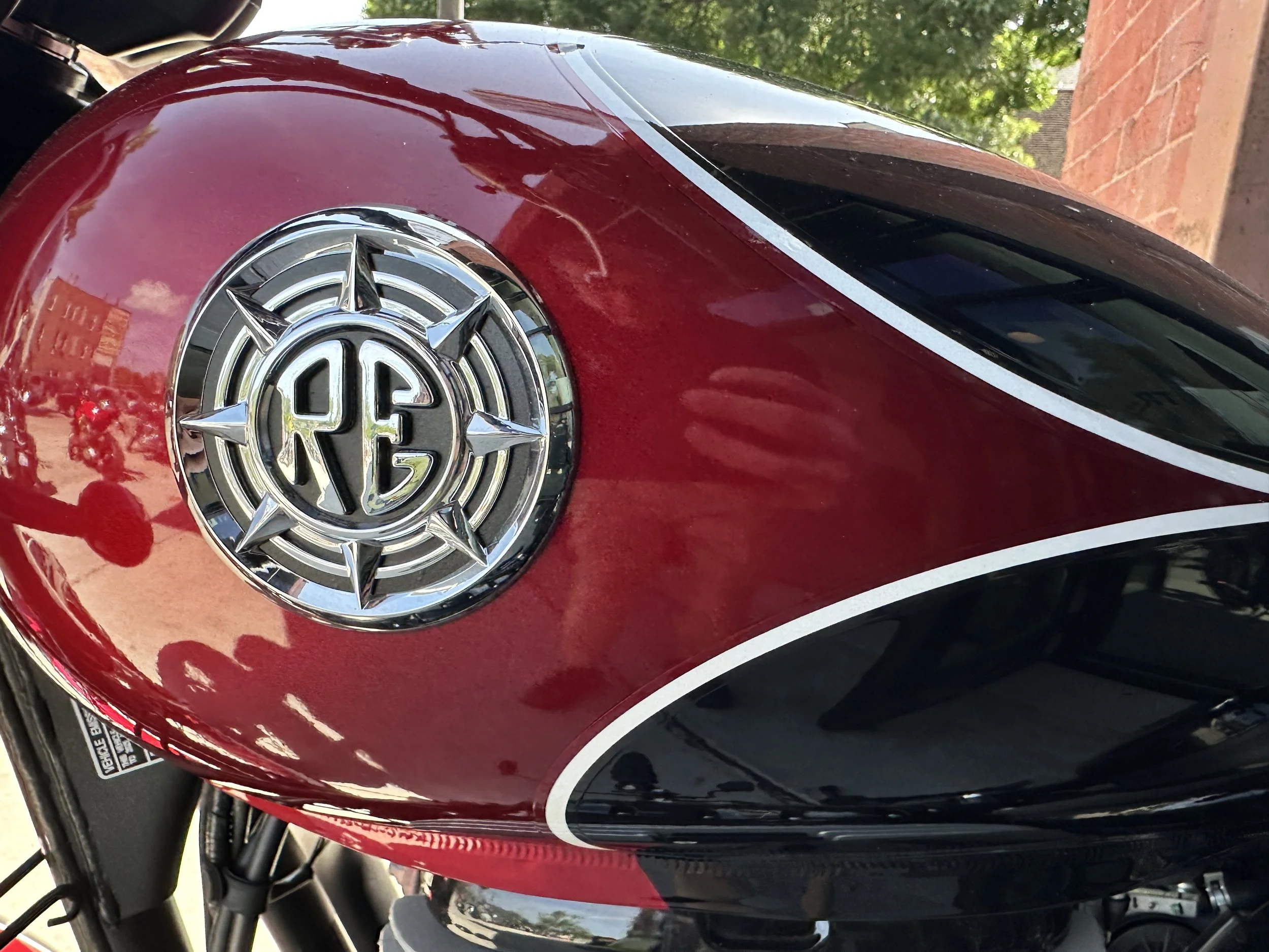 Close-up of a red and black motorcycle with a chrome emblem on the fuel tank, reflecting trees and the sky.