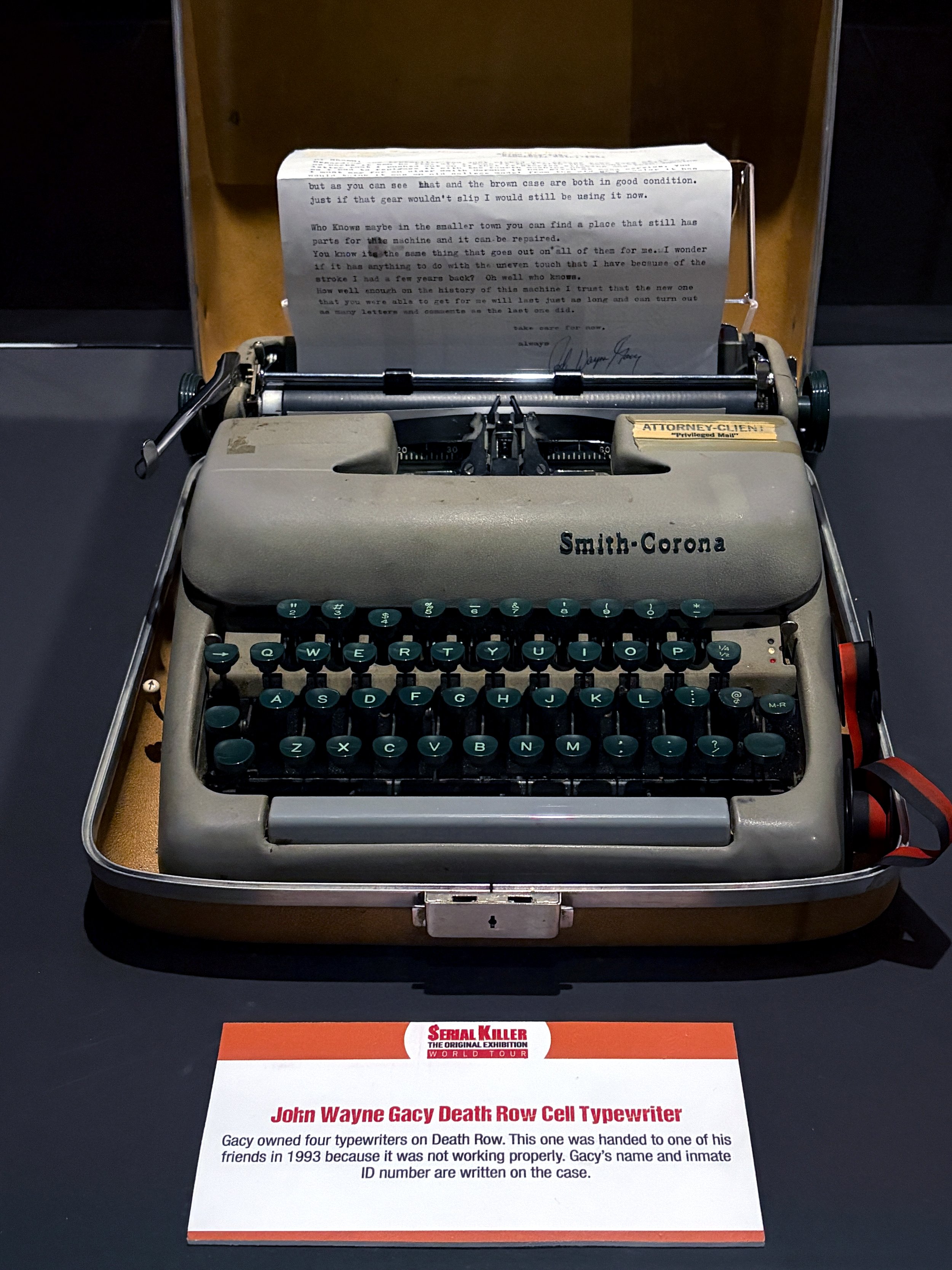 A vintage Smith-Corona typewriter with a sheet of paper inside. In front of it, there is a sign that reads: 'John Wayne Gacy Death Row Cell Typewriter. Gacy owned four typewriters on Death Row. This one was handed to one of his friends in 1993 becaus