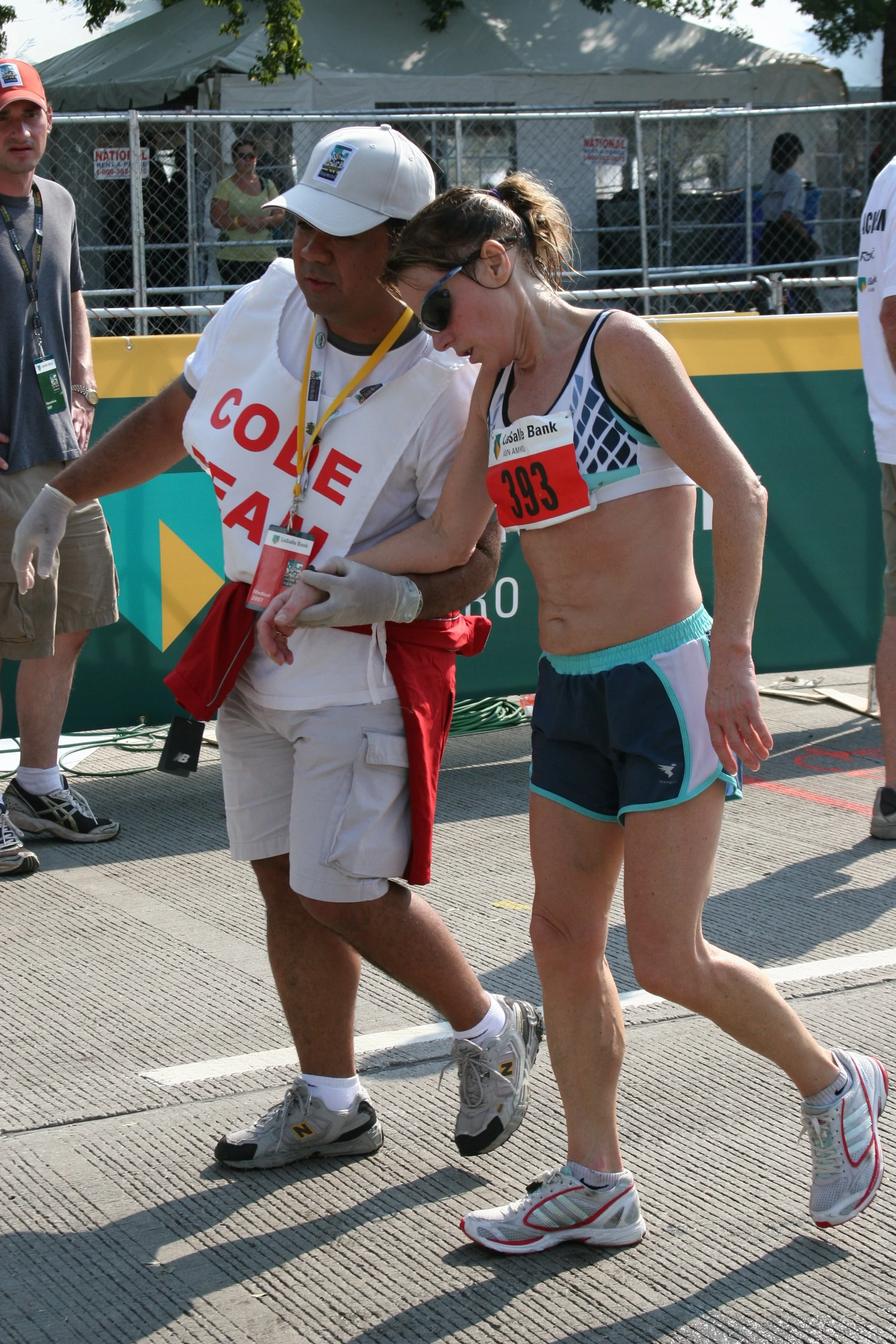 A female marathon runner wearing sunglasses, a sports top, and shorts, being attended by a male medical worker dressed in a white vest with red text and white shorts during a race event, with a green and yellow barricade and chain-link fence in the b