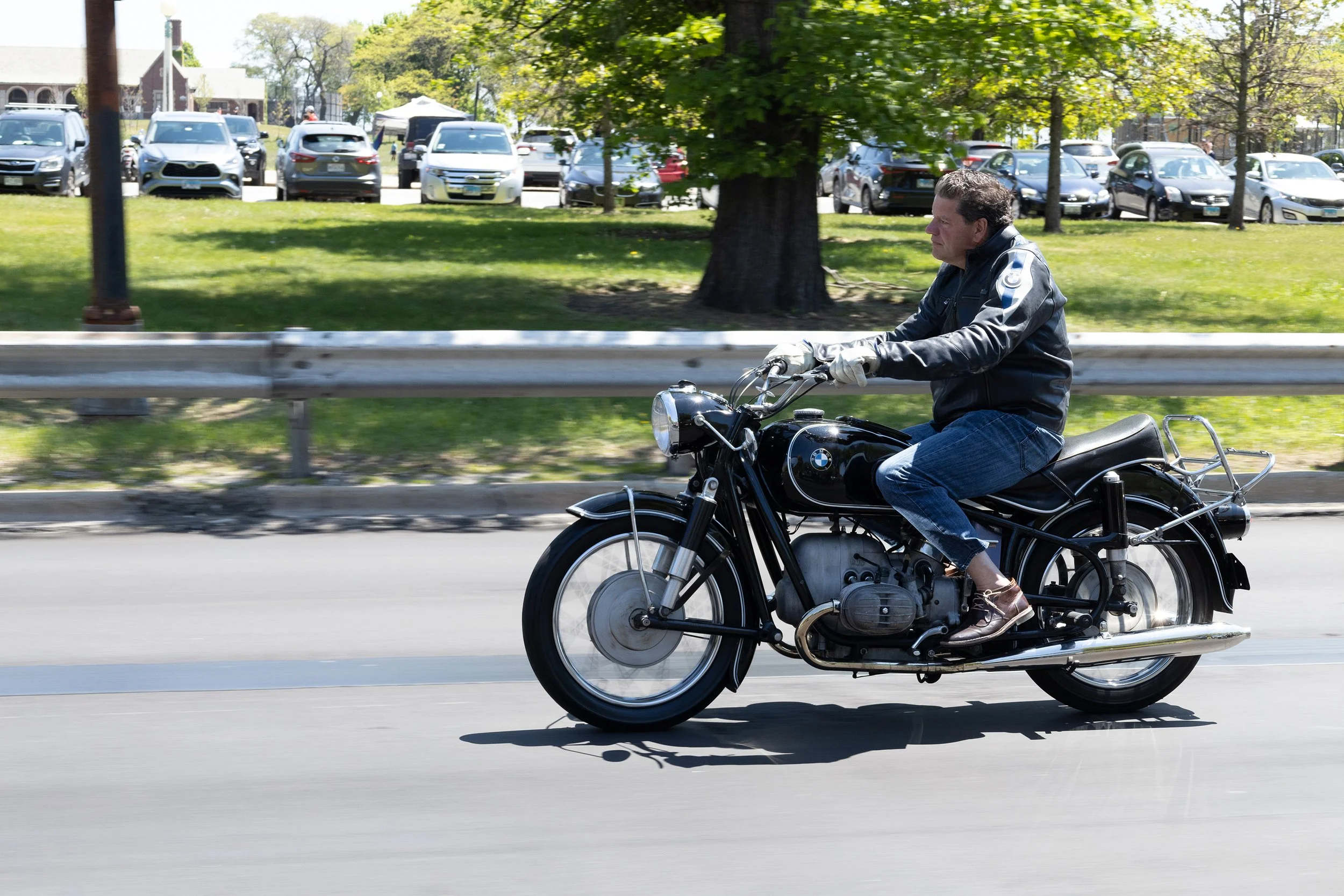 A man riding a black motorcycle on a city street with a background of parked cars and green trees.