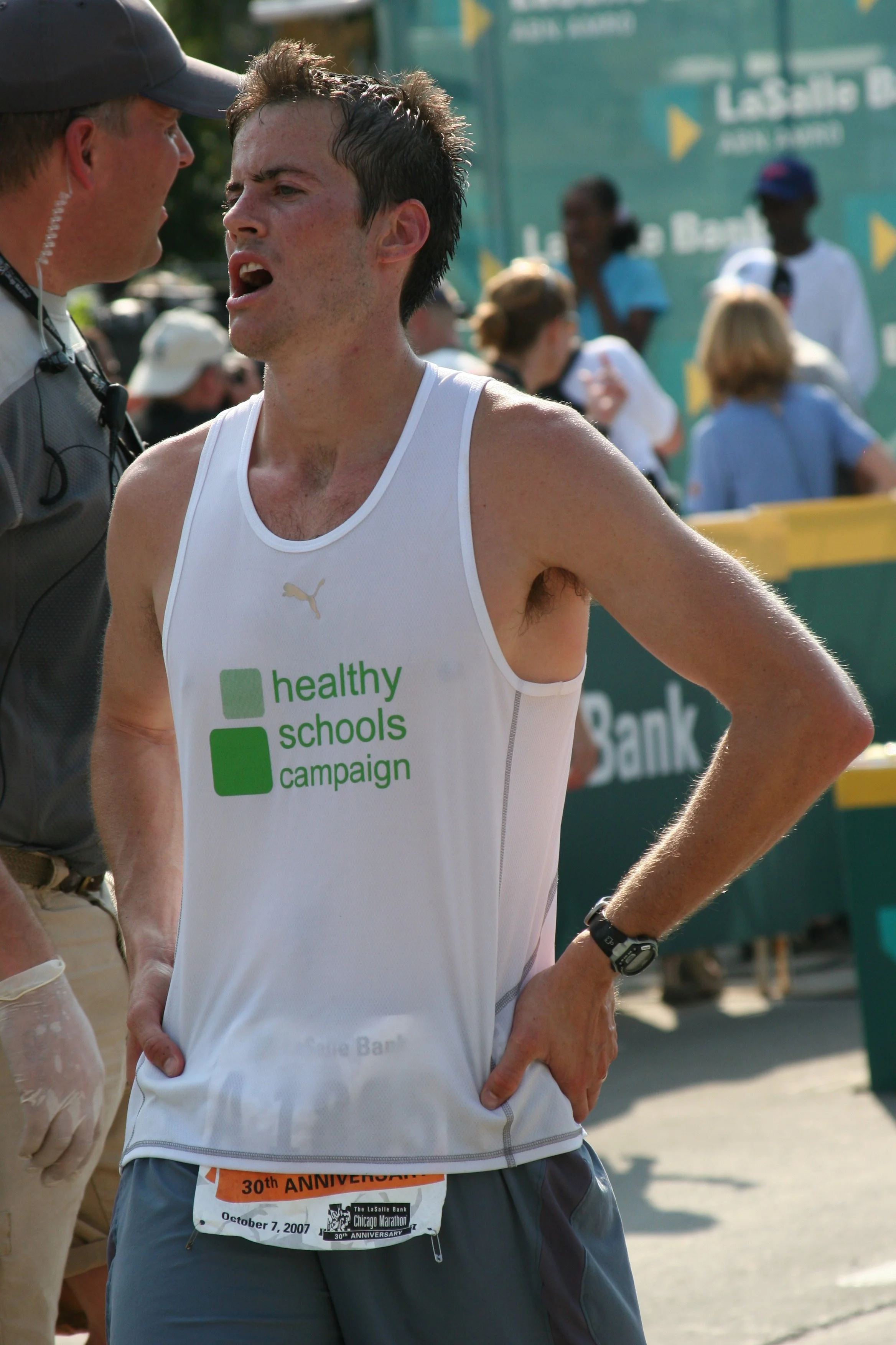 A young man wearing a white running shirt with 'healthy schools campaign' printed on it, standing outdoors at a marathon event, speaking to a race official. He has dark, wet hair and a race bib showing it is the 30th anniversary of the Chicago Marath