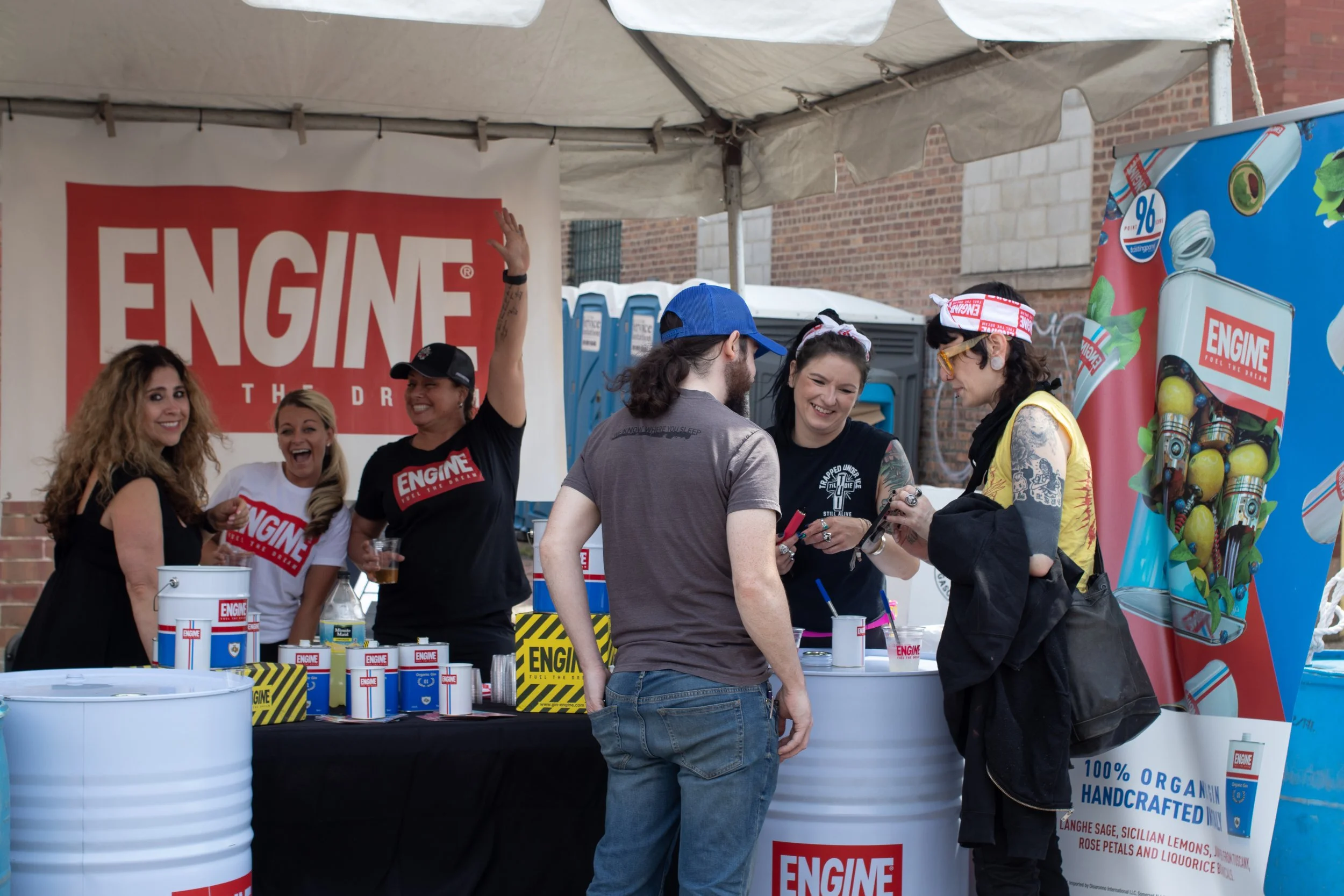 A group of people gathers at an outdoor booth promoting engine products, with a large red and white sign reading 'ENGINE'. They are smiling and engaged in conversation, with promotional items and cans labeled 'ENGINE' on the table.