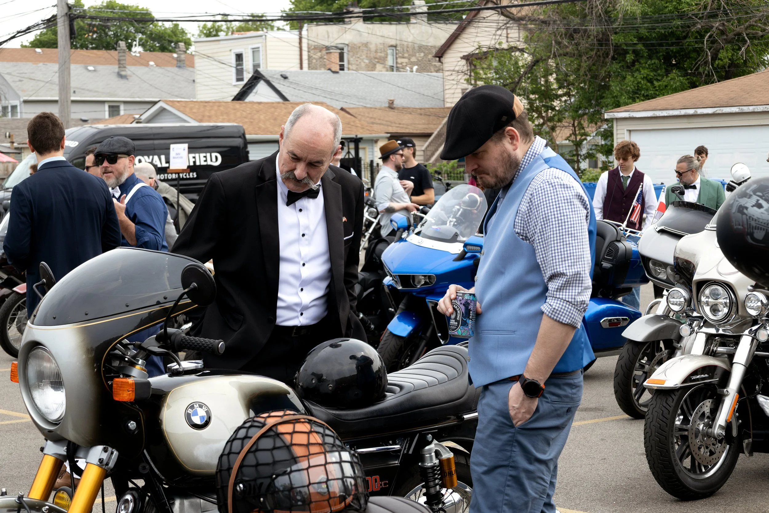 A group of people gathered around motorcycles in an outdoor parking lot, with two men in the foreground inspecting a BMW motorcycle and others in the background engaging with each other.
