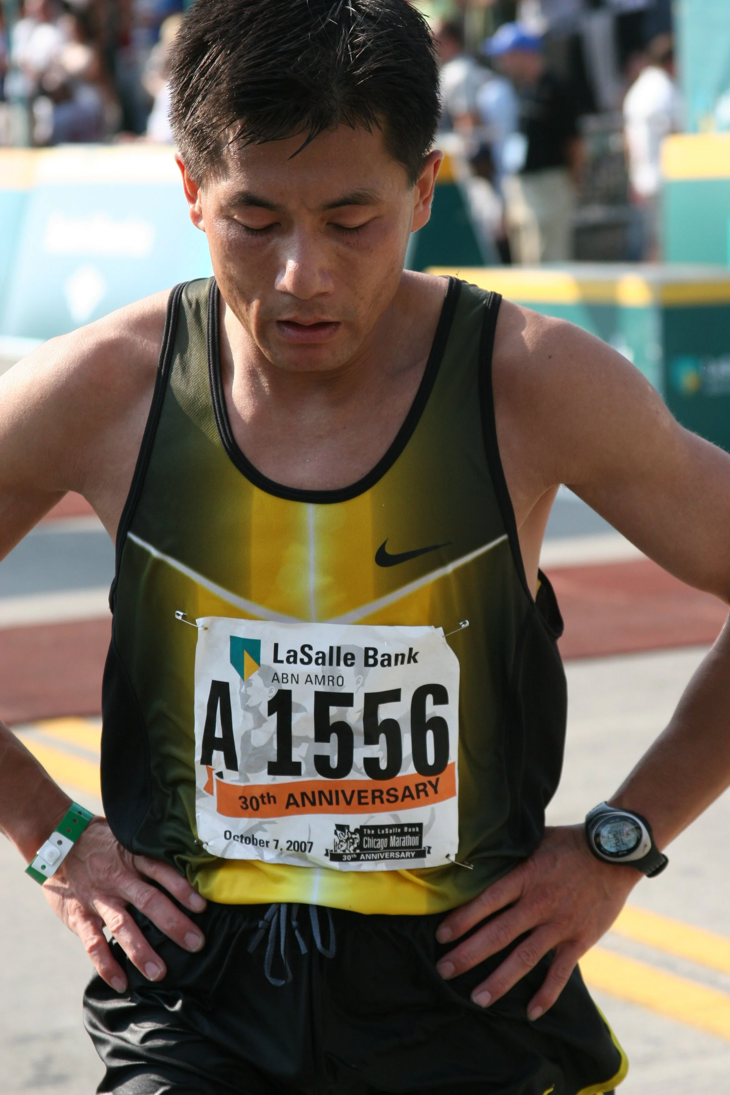 A male marathon runner wearing a yellow and black athletic shirt, with a bib number A1556. The bib indicates the 30th anniversary of the Chicago Marathon on October 7, 2007. He appears exhausted, with his eyes closed and a facial expression of fatigu
