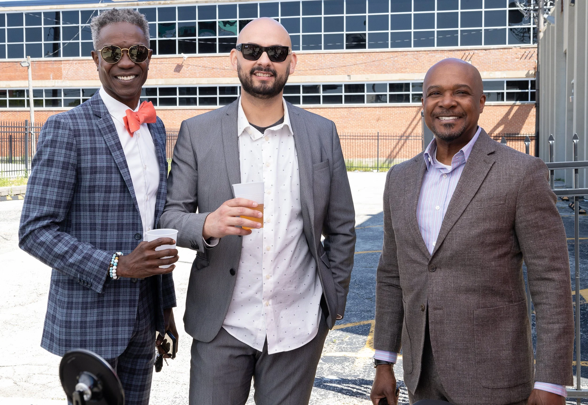 Three men dressed in suits and sunglasses standing outdoors, smiling at the camera, with a building and fencing in the background.
