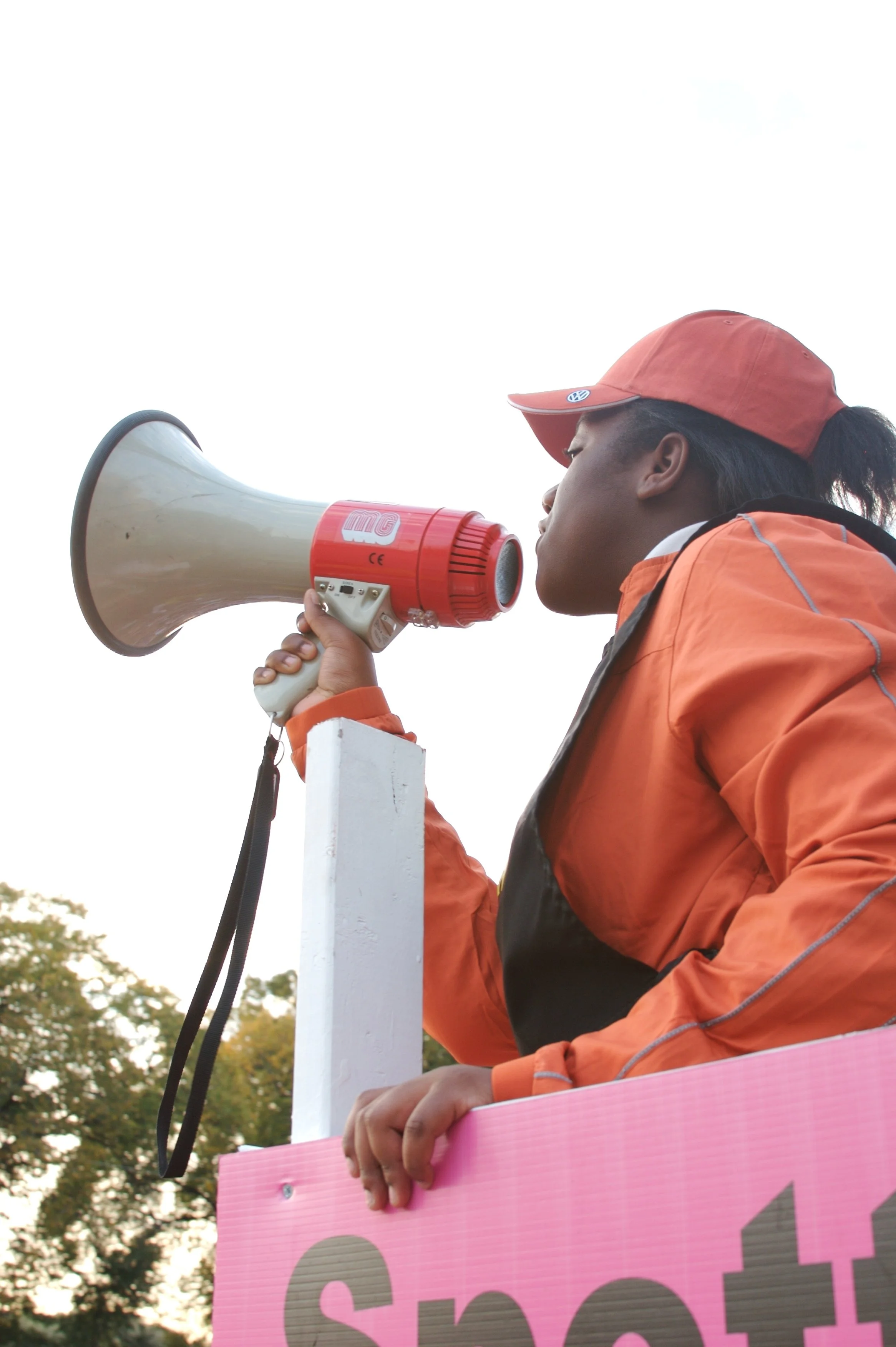 Woman in orange jacket and red cap speaking into a megaphone during a protest or rally, holding a pink sign.