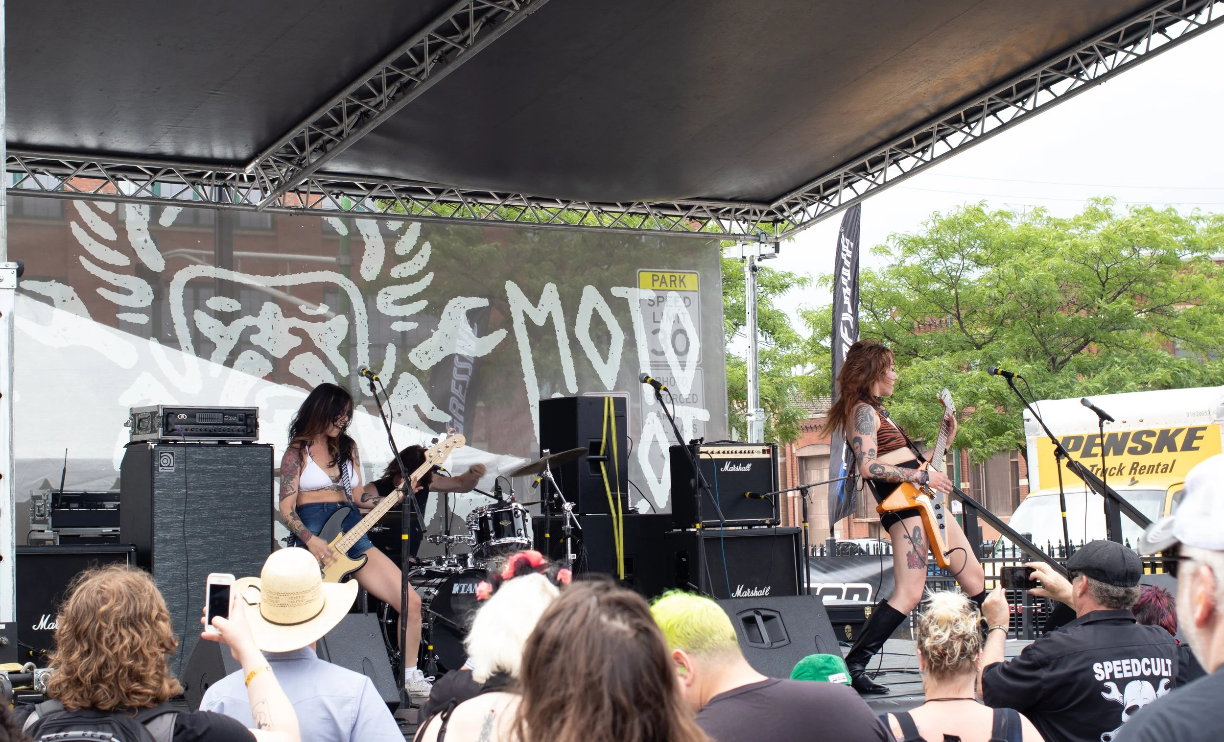 A live outdoor band performance on a stage with three female musicians, one playing bass, one on drums, and one with an electric guitar, during a festival. Audience members watching, some taking photos. The stage has musical equipment including ampli