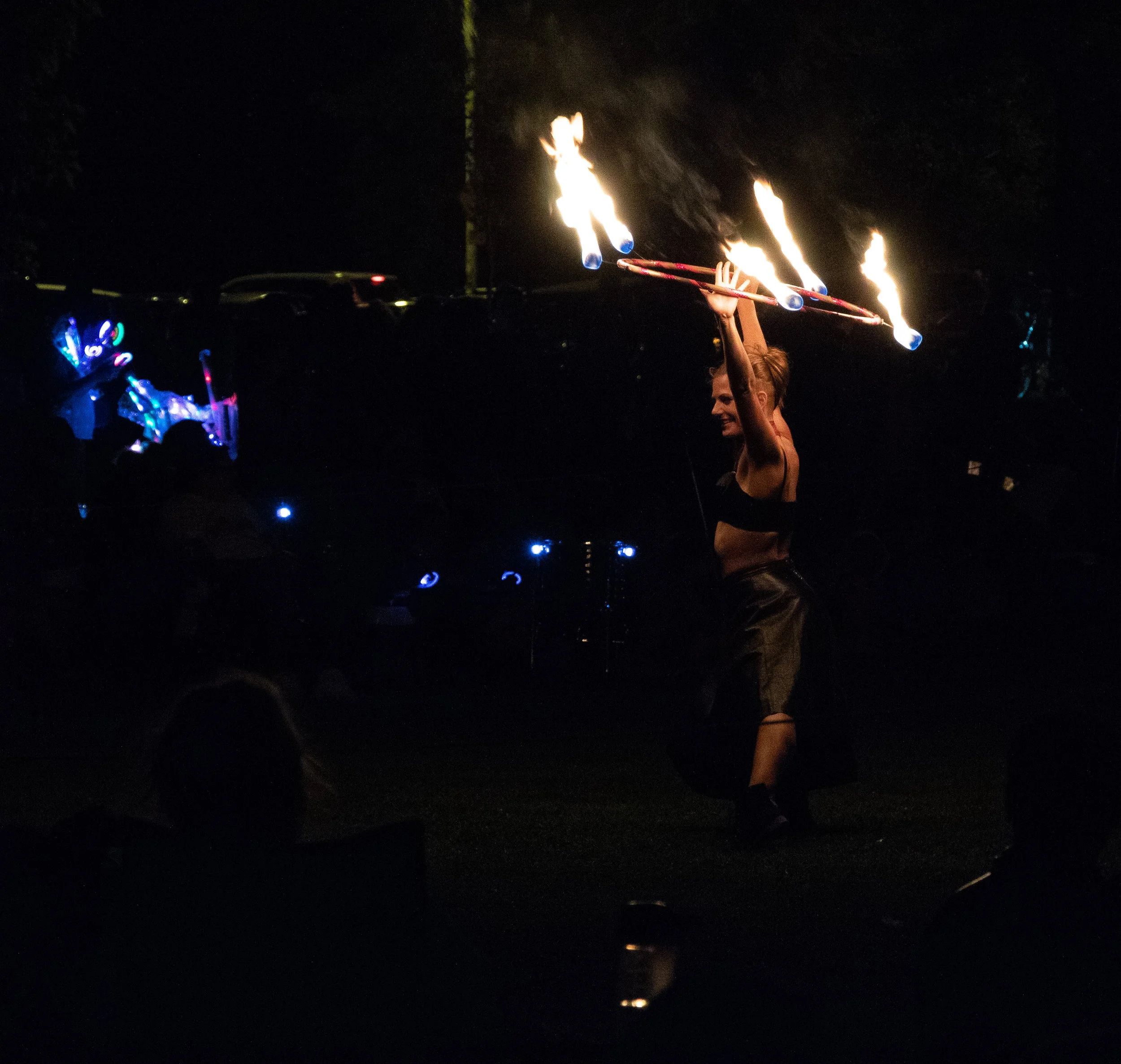 A person holding flaming poi sticks at night, with the dark sky and some faintly visible cars in the background.