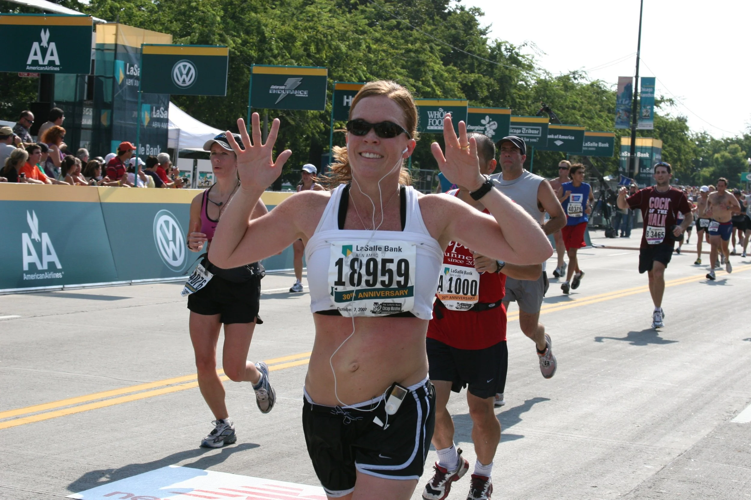 Female marathon runner smiling at the camera while participating in a race on a sunny day. Other runners and spectators are visible in the background, and race banners and signs line the street.