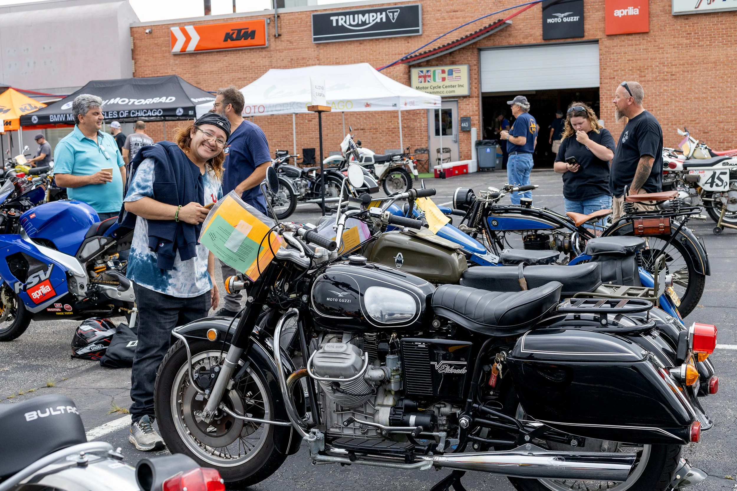 A group of people at a motorcycle gathering outside a brick building with various motorcycle brand signs. Several vintage and modern motorcycles are displayed, and some individuals are engaged in conversation, looking at the bikes, or using their sma