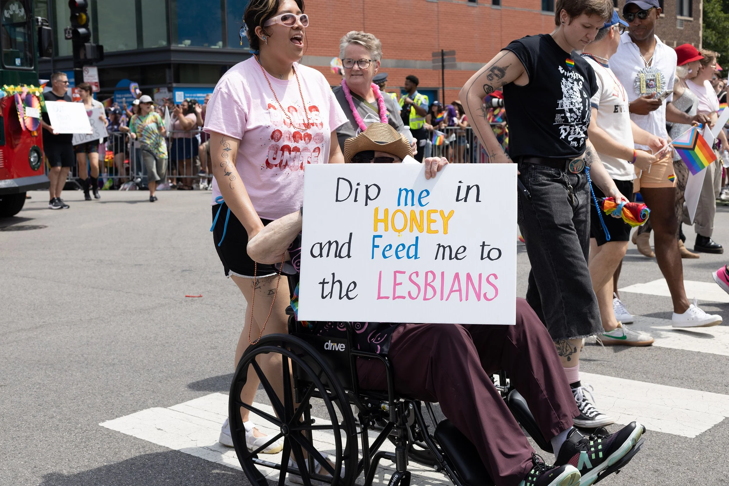 Woman in a pink T-shirt standing beside a person in a wheelchair holding a sign that reads, 'Dip me in honey and feed me to the lesbians,' during a pride parade. The scene is filled with people carrying rainbow flags and signs, with a red brick build