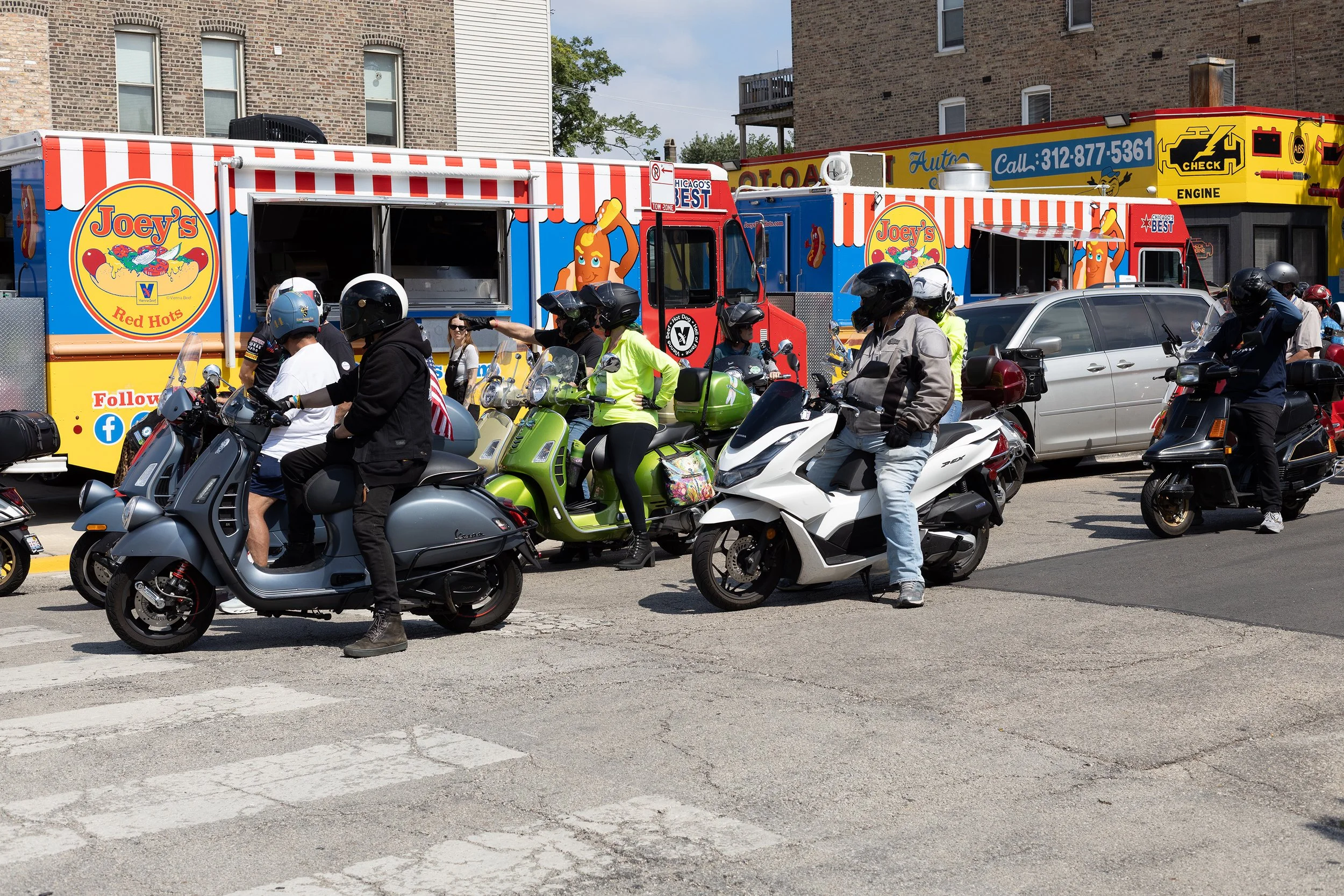 Multiple motorcyclists and scooters are gathered at an intersection, with a food truck named Joey's Red Hots in the background. Several people are wearing helmets, and there are buildings lining the street.