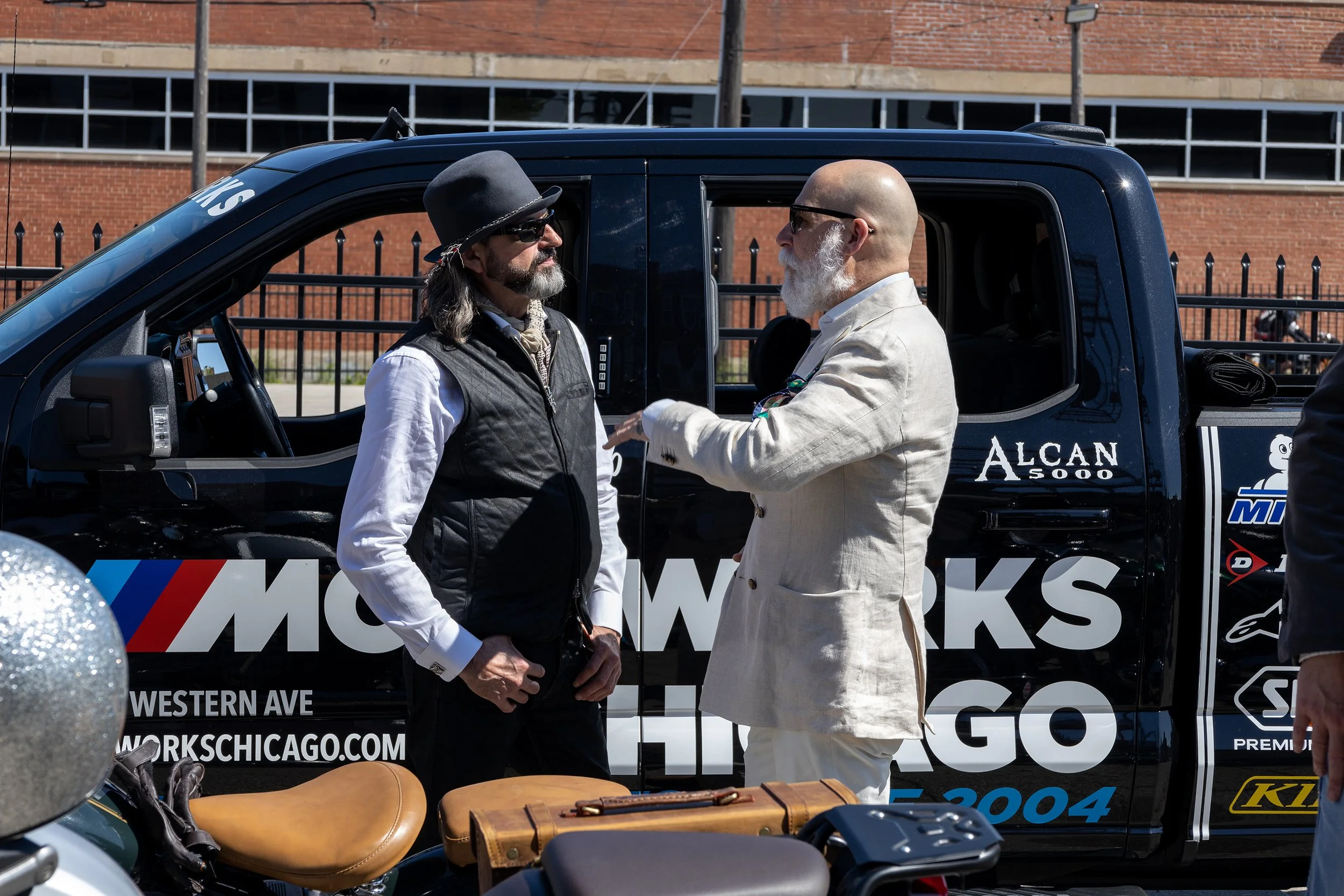 Two men with beards and sunglasses are having a conversation next to a black vehicle with promotional signage on it, in an outdoor urban setting.