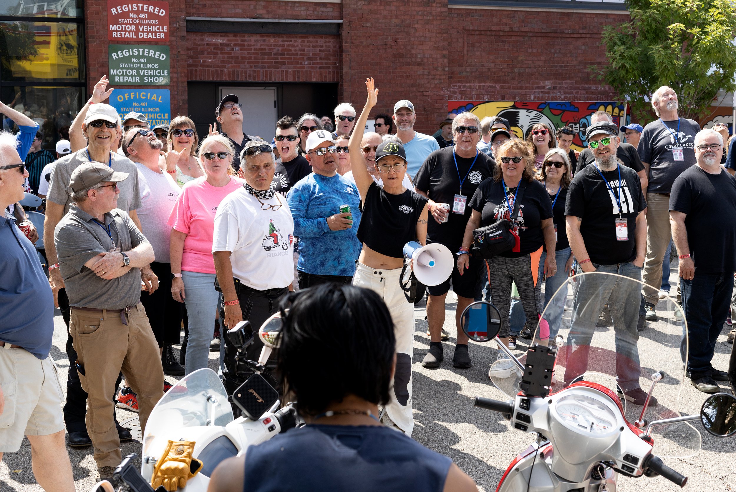 A large group of people gathered outside a brick building, some wearing sunglasses, holding drinks, and smiling, with motorcycles parked in the foreground.