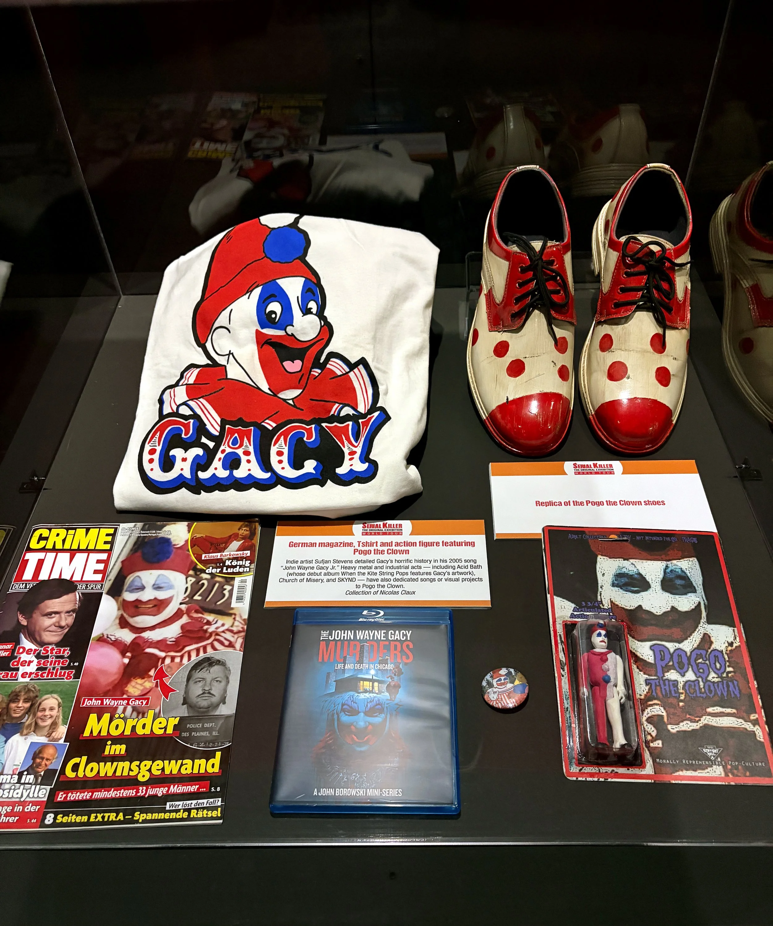 Display case with various horror-themed items including a book titled 'Murder in Clownland,' a mini-series DVD of John Borowski's 'Gacy,' a book about John Wayne Gacy, a model of Pogo the Clown, and a pair of clown shoes with red polka dots.