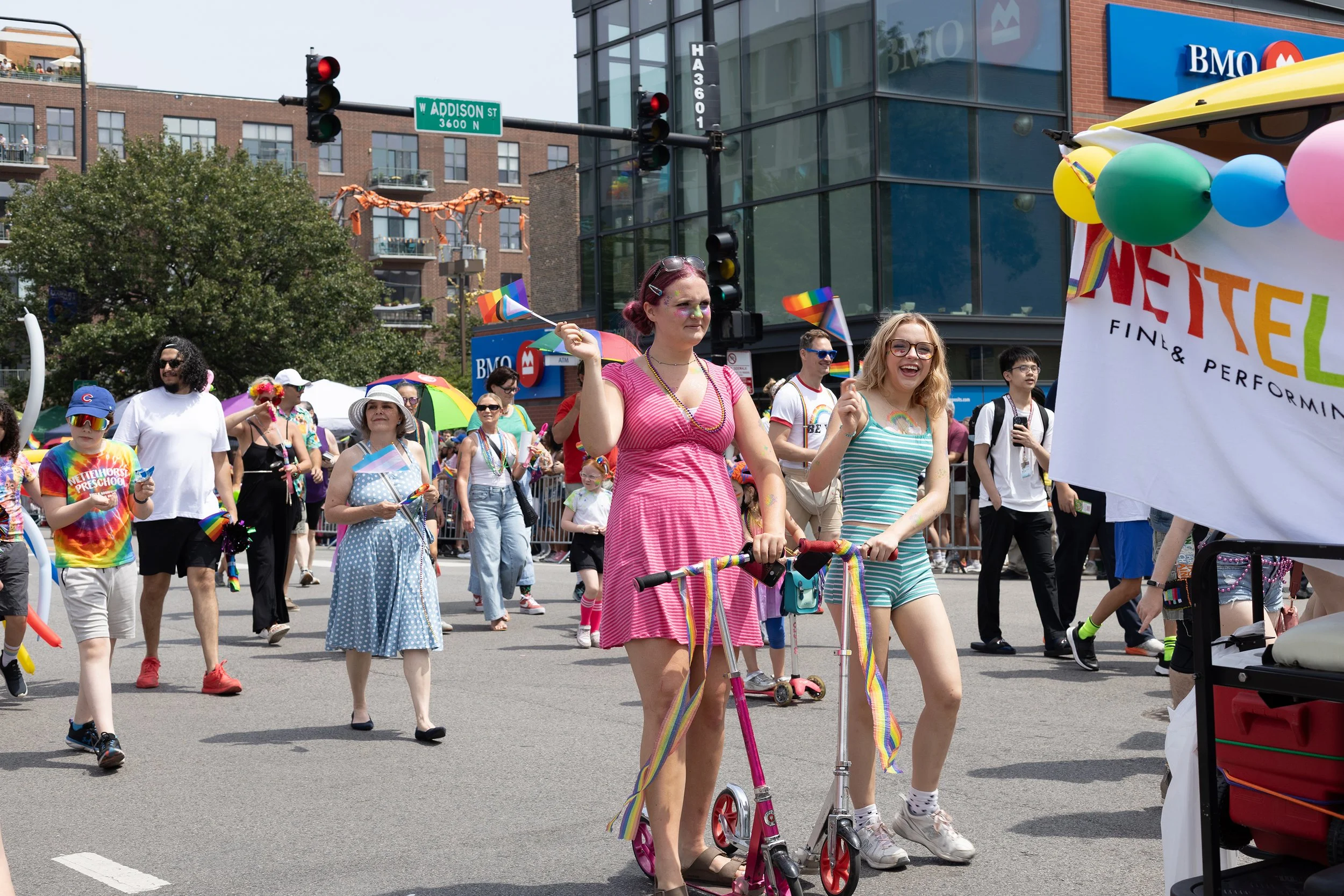 People participating in a pride parade, holding rainbow flags, walking on the street in front of buildings with a BMO bank sign. Some individuals are wearing colorful clothing and accessories, with a festive atmosphere.