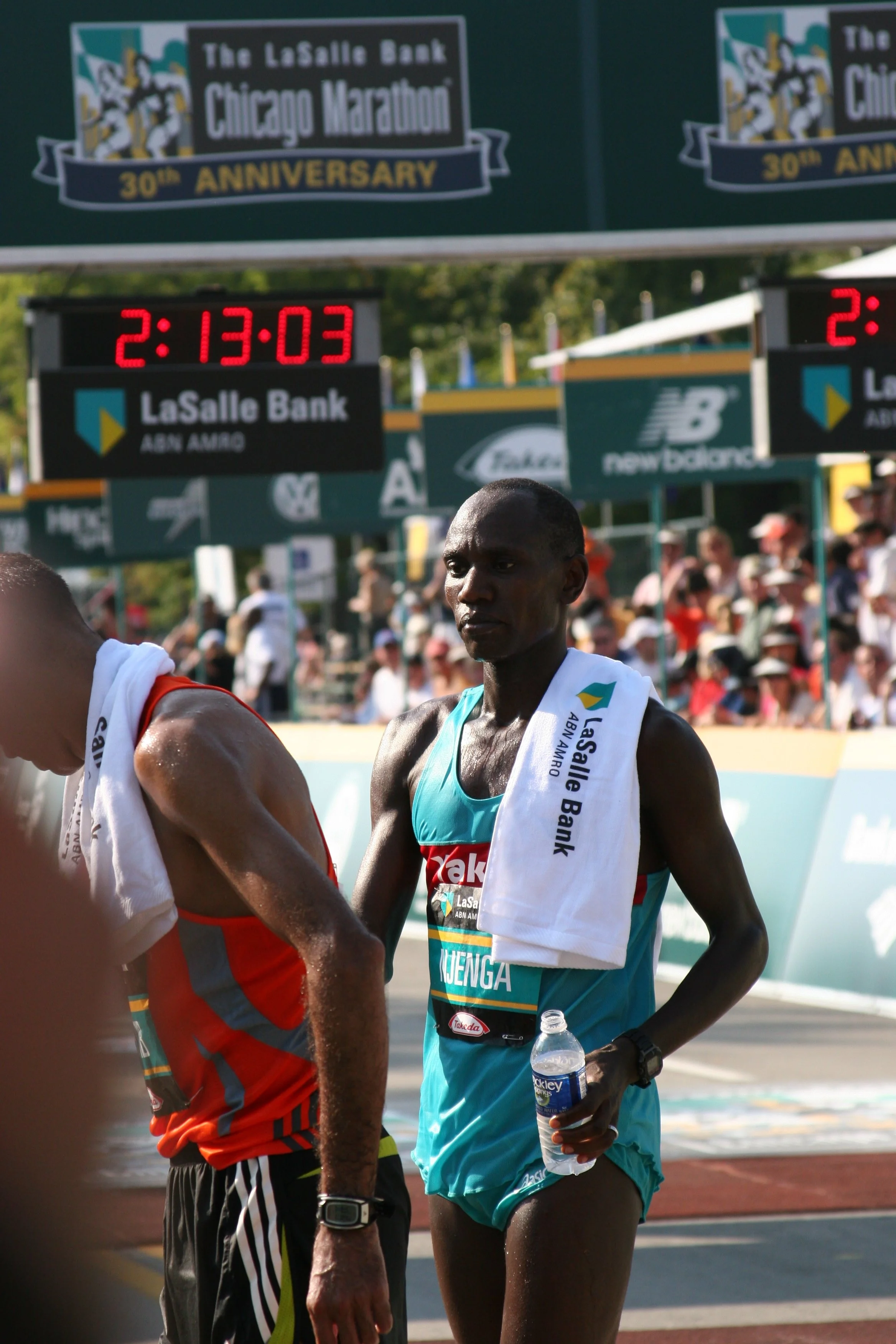 Marathon runners at the finish line of the Chicago Marathon, with a digital clock showing 2:13:03, and a banner indicating the event's 30th anniversary.