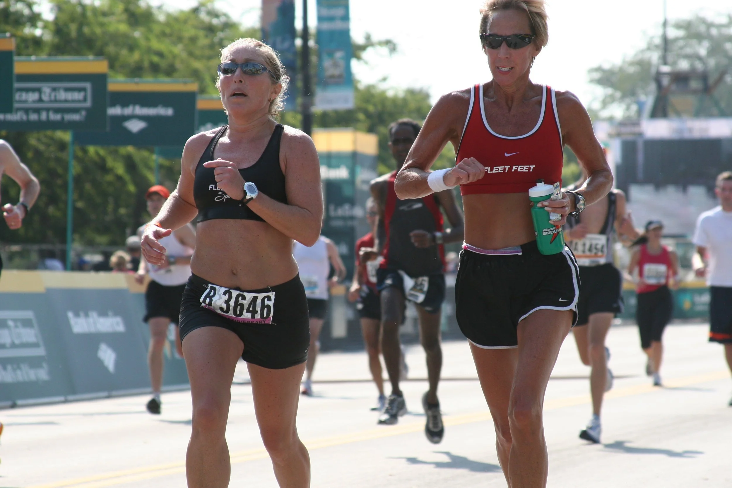 Two women running in a race on a city street with other runners in the background, wearing athletic clothing and sunglasses.