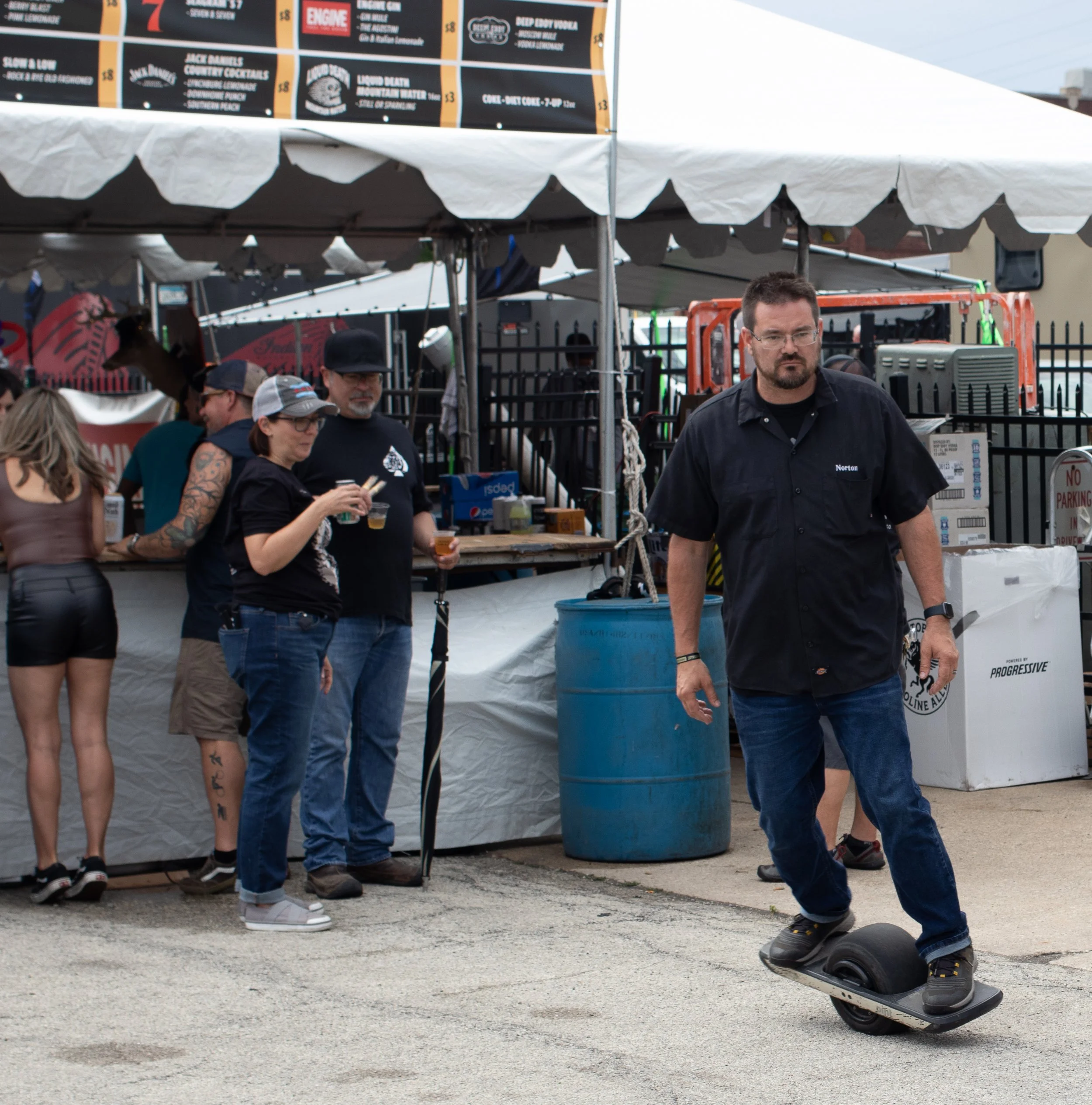 People standing and walking at an outdoor event or festival with a white tent, food and drink stalls, and a person riding a hoverboard.