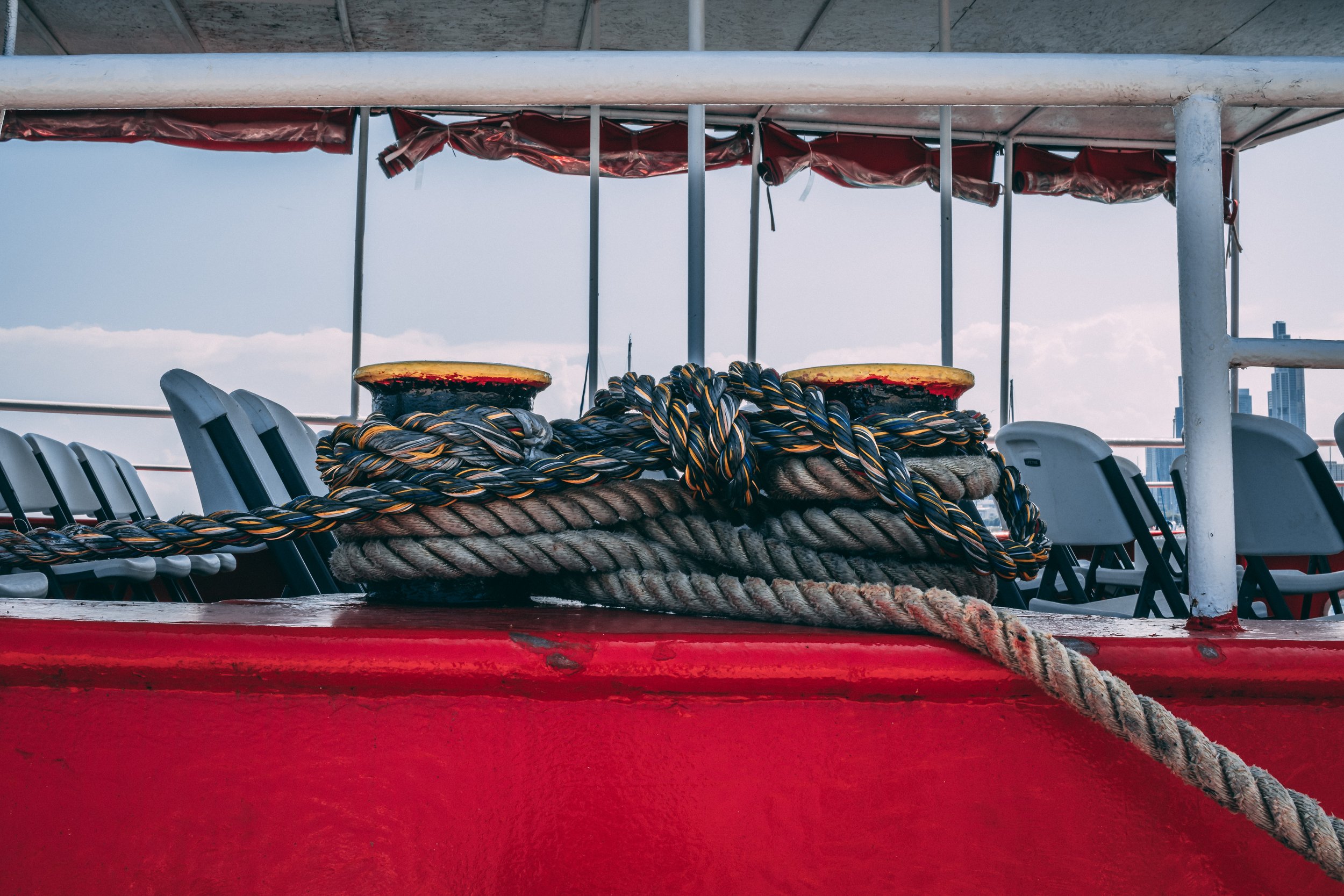 Close-up of a ship's deck with thick ropes coiled around a winch, set against a background of chairs, structure, and a city skyline in the distance.