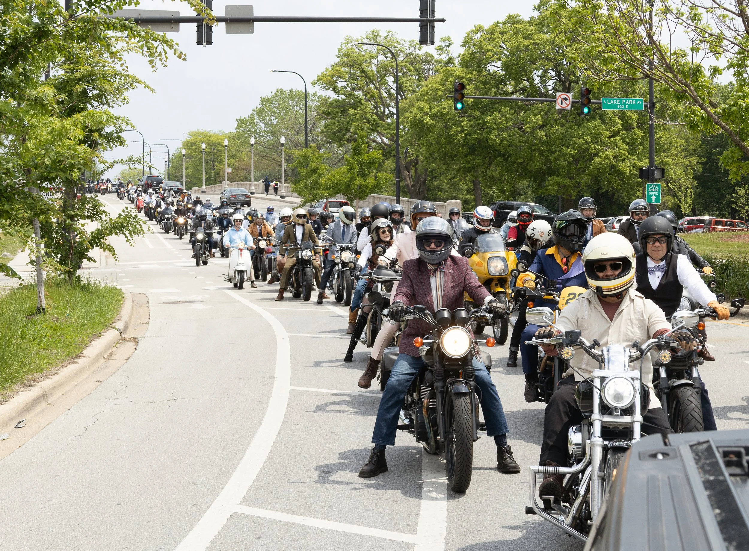 A large group of motorcyclists riding their bikes on a city street, passing underneath traffic lights and surrounded by green trees.