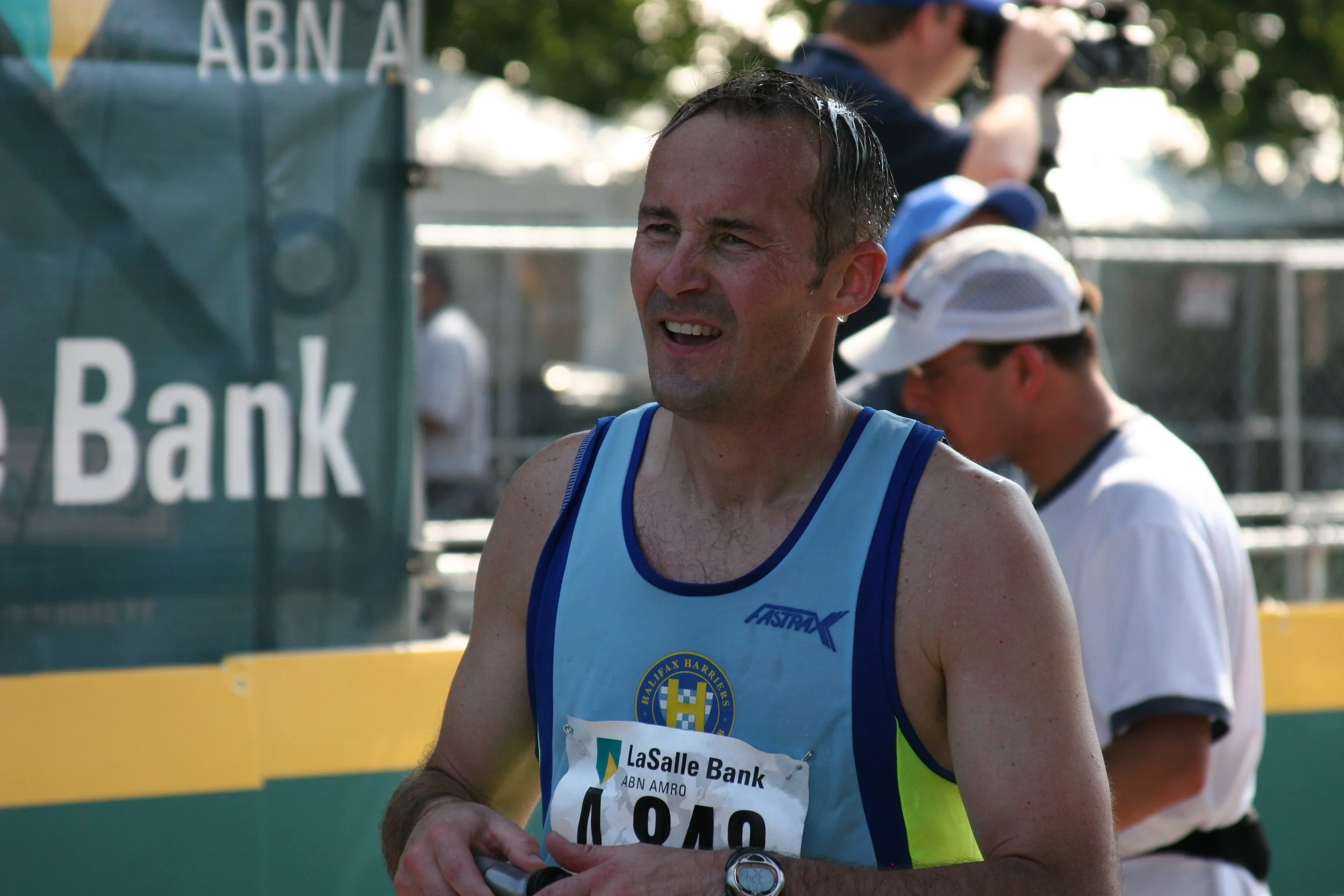 A male marathon runner wearing a blue tank top with the logo of Halifax Harriers, holding a device in his hands during a race. Behind him is a spectator with a camera and another person with a cap, all outdoors with trees and a sign partially visible