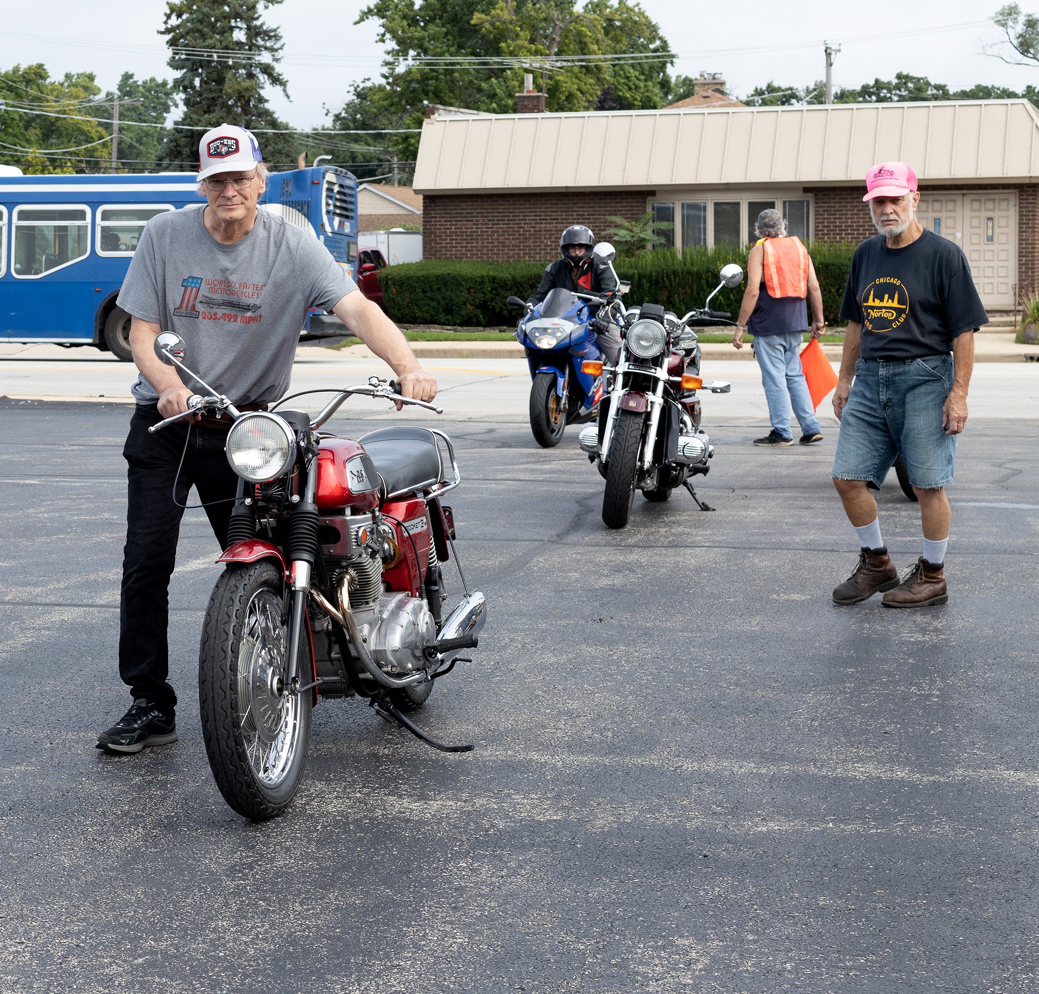 A group of people gathered outdoors with motorcycles and a scooter in front of houses. Two older men are in the foreground, one on a scooter wearing a gray t-shirt and cap, the other standing beside a motorcycle wearing a black t-shirt and pink cap. 