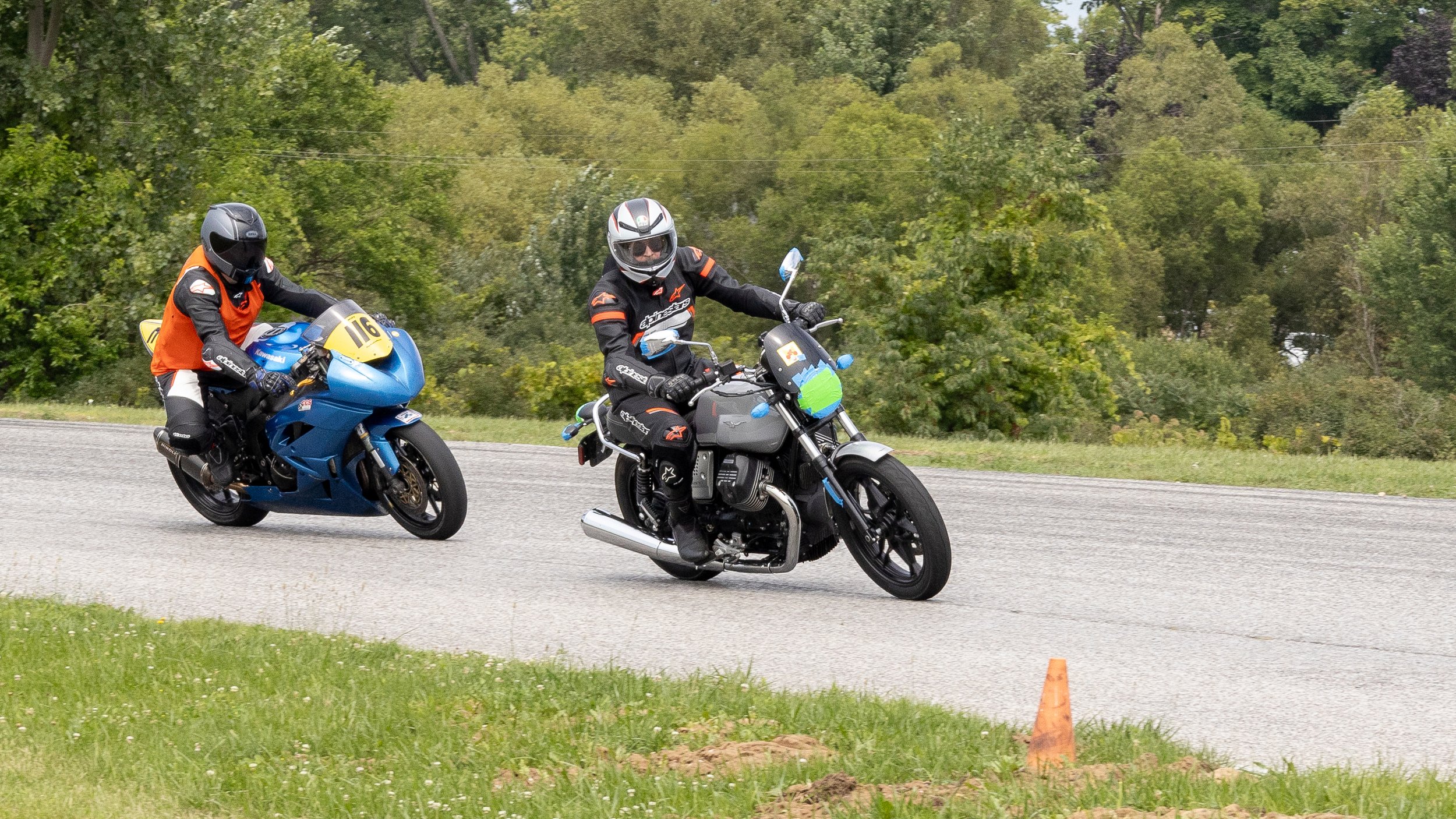 Two motorcyclists riding on a road bordered by trees, with orange cones on the side.